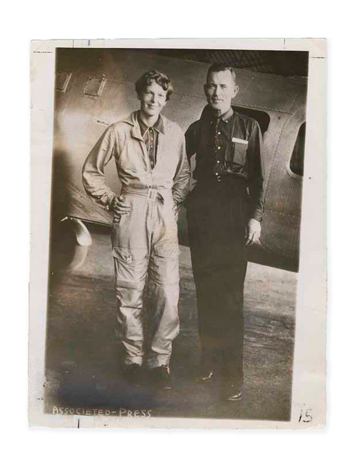 A black and white photo of a Amelia Earhart in a jumpsuit and Fred Noonan in a military uniform standing in front of an airplane.