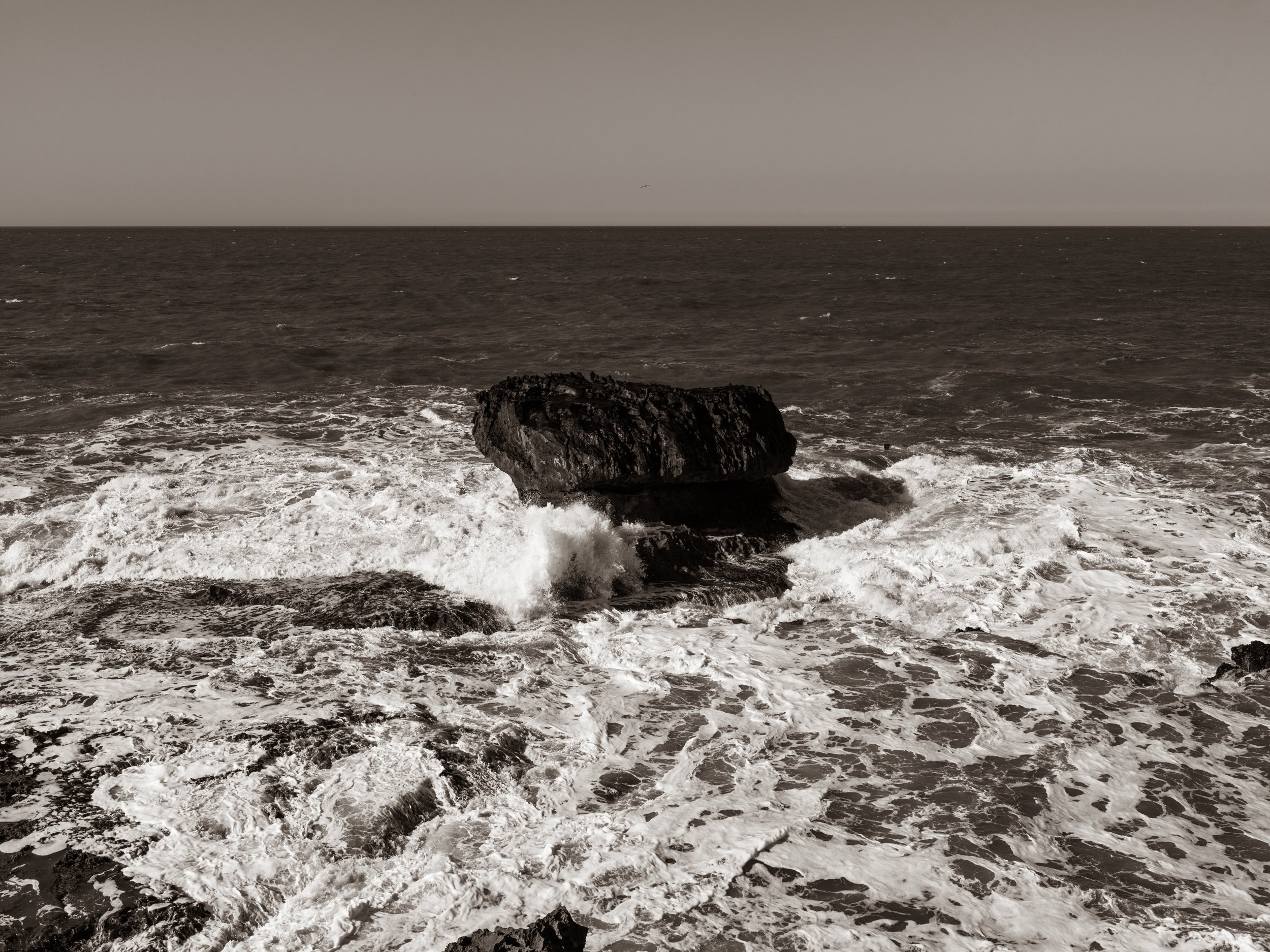 Dark ocean water with white foamy waves crashing near rocks, under a cloudy sky