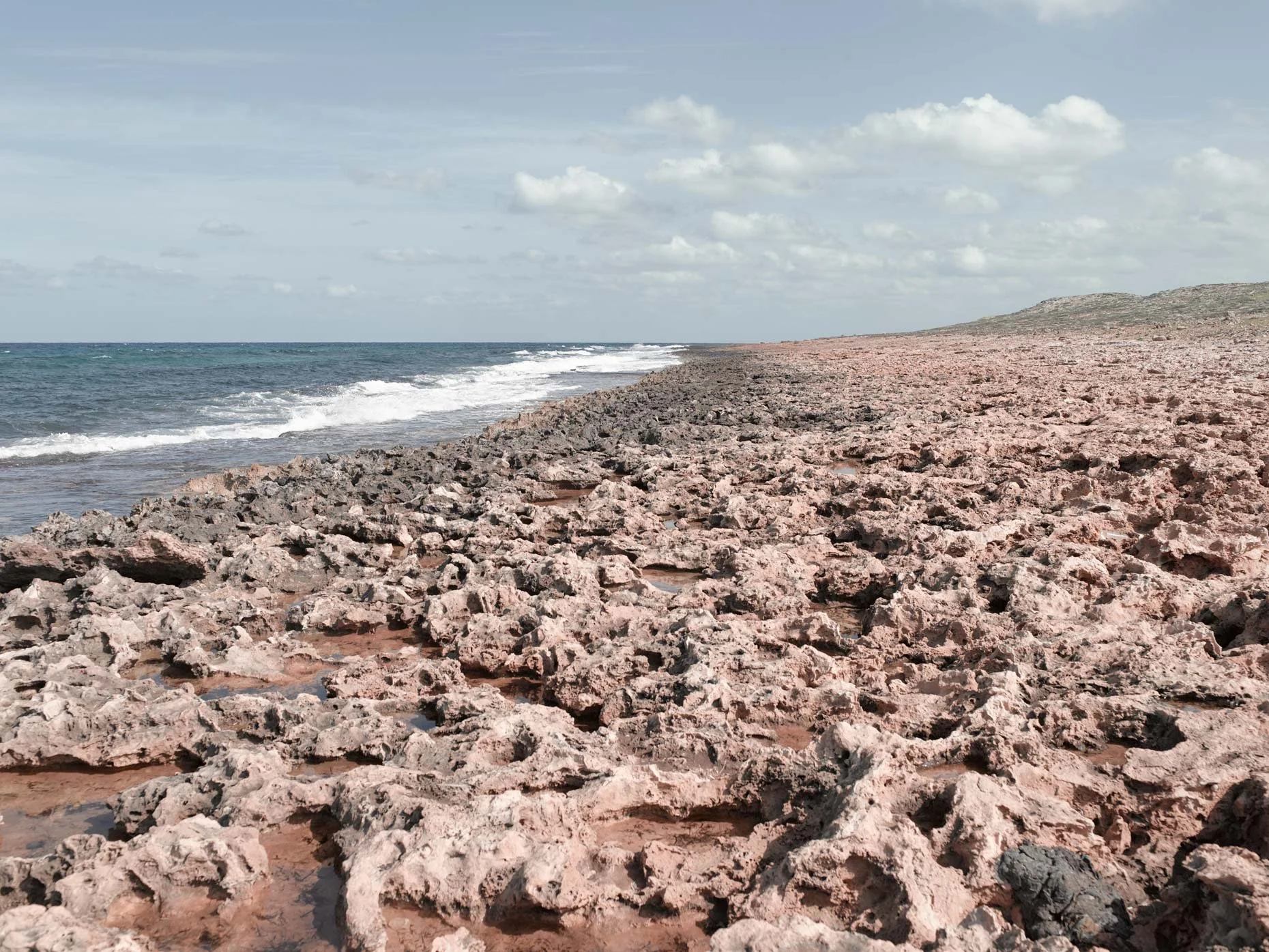 Rocky beach shoreline with ocean waves, distant hill, and cloudy sky.