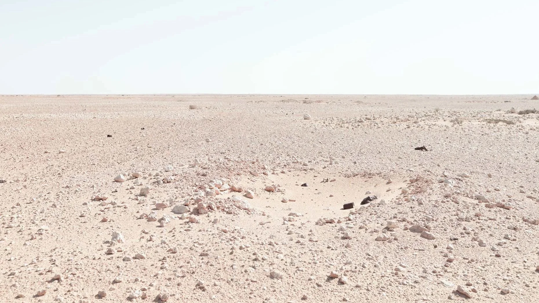 A wide view of a flat, barren desert landscape with sparse rocks and a pale, sandy surface under a clear sky.