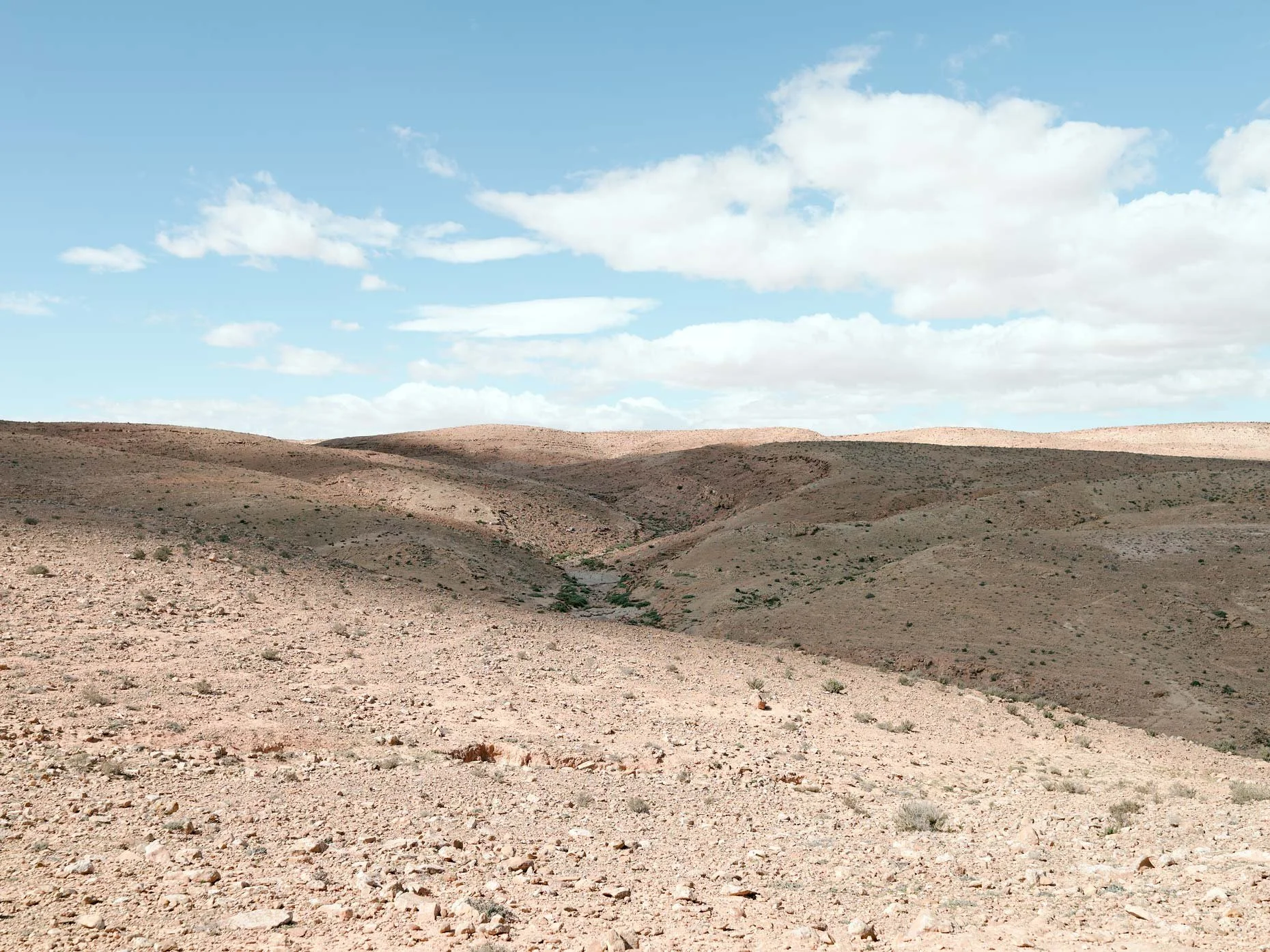 A desert landscape with rocky, sandy terrain and sparse vegetation, rolling hills in the distance, under a bright blue sky with scattered white clouds.