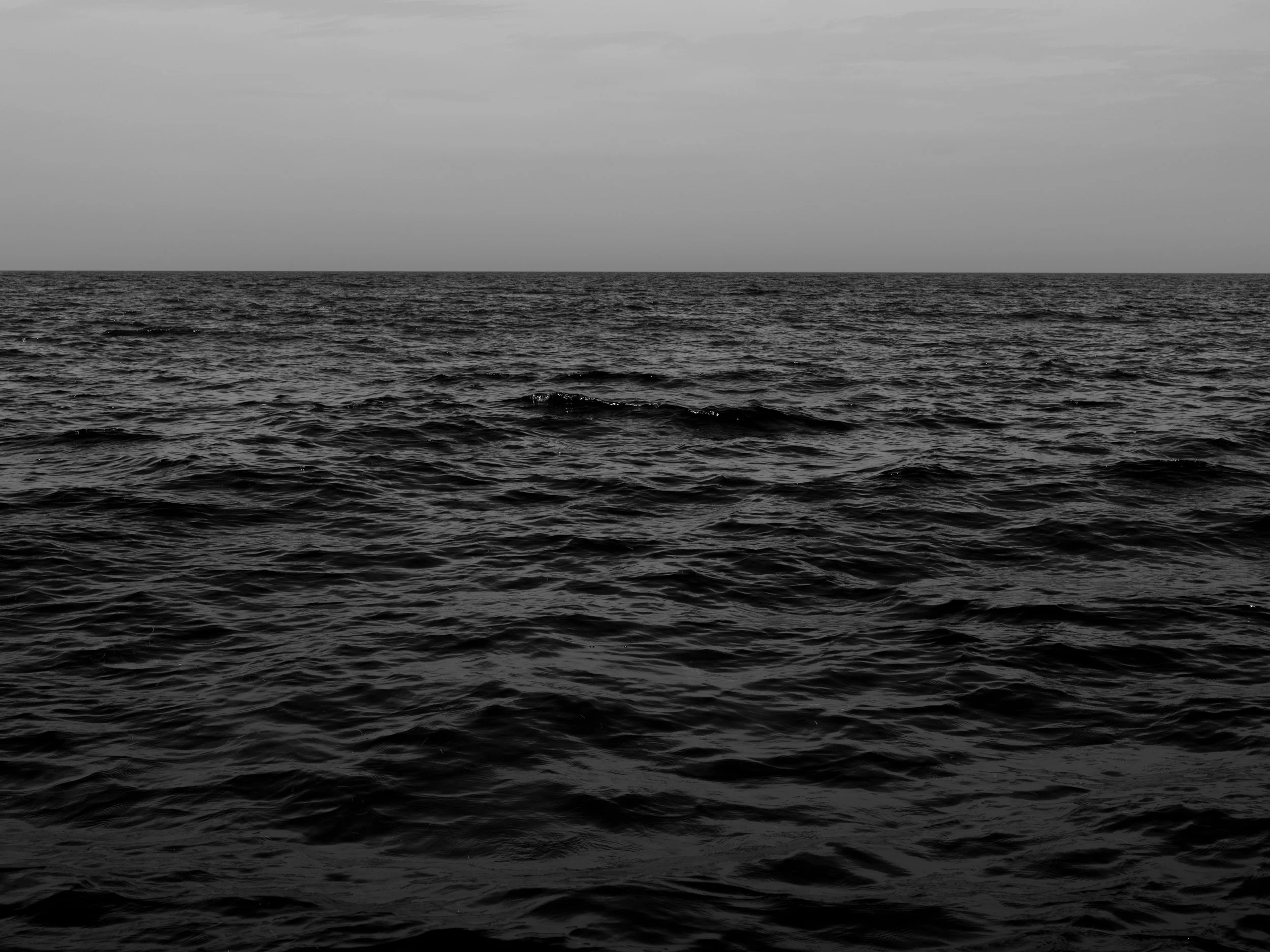 A black and white photograph of the ocean with calm water and a cloudy sky.