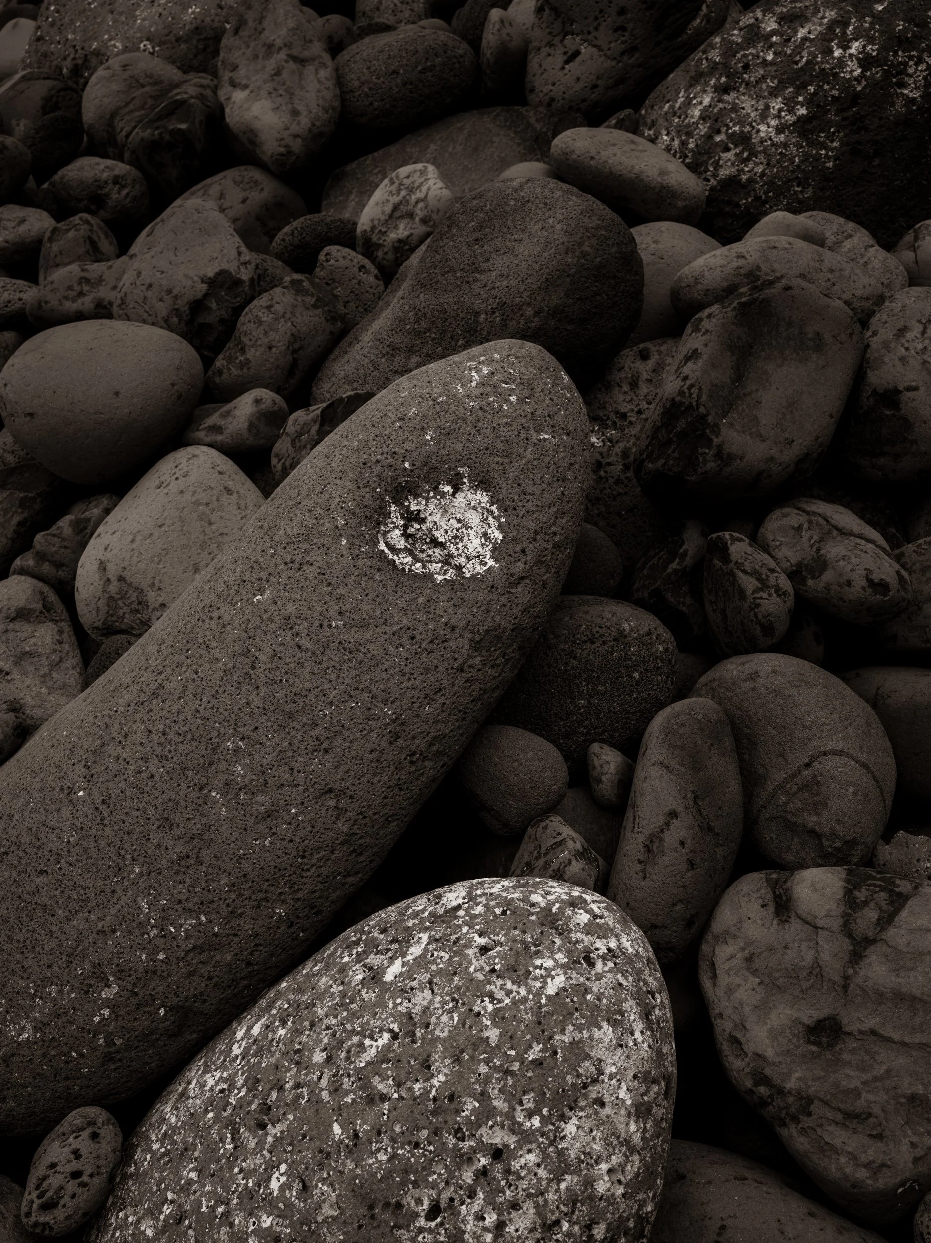 A collection of smooth, rounded gray and dark stone rocks of various sizes, some with patches of white lichen or mineral deposits.