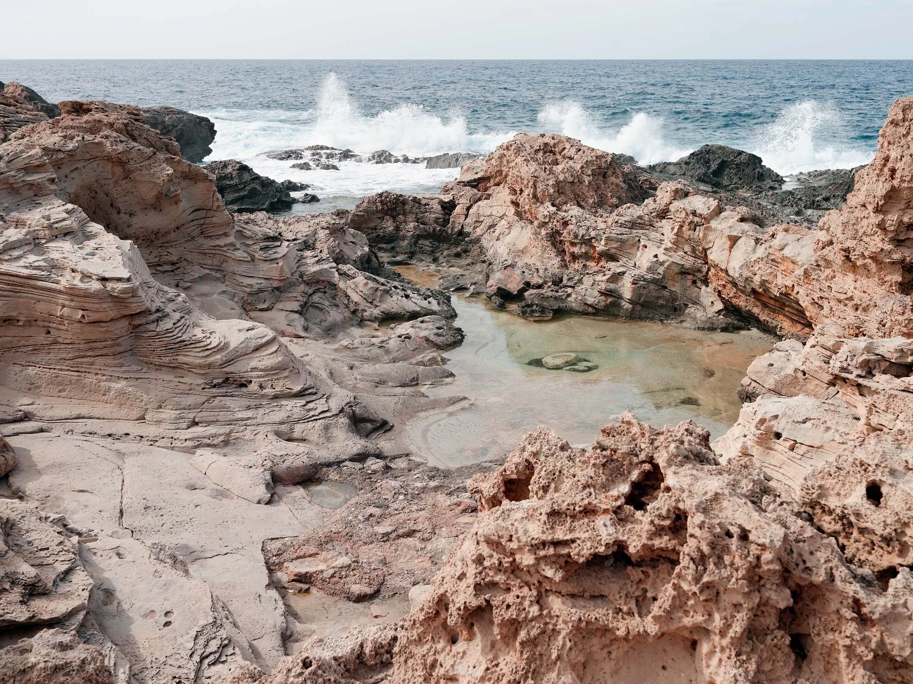 A rocky coastline with beige and reddish rocks and a small pool of water, with ocean waves crashing against the rocks in the background.