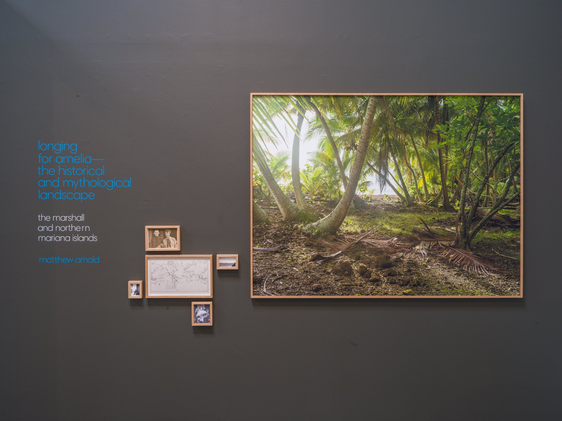 Exhibition display featuring a large framed photograph of a tropical landscape with palm trees and greenery, accompanied by a text panel on the left that references the historical and mythological landscape of the Marshall and northern Mariana Island