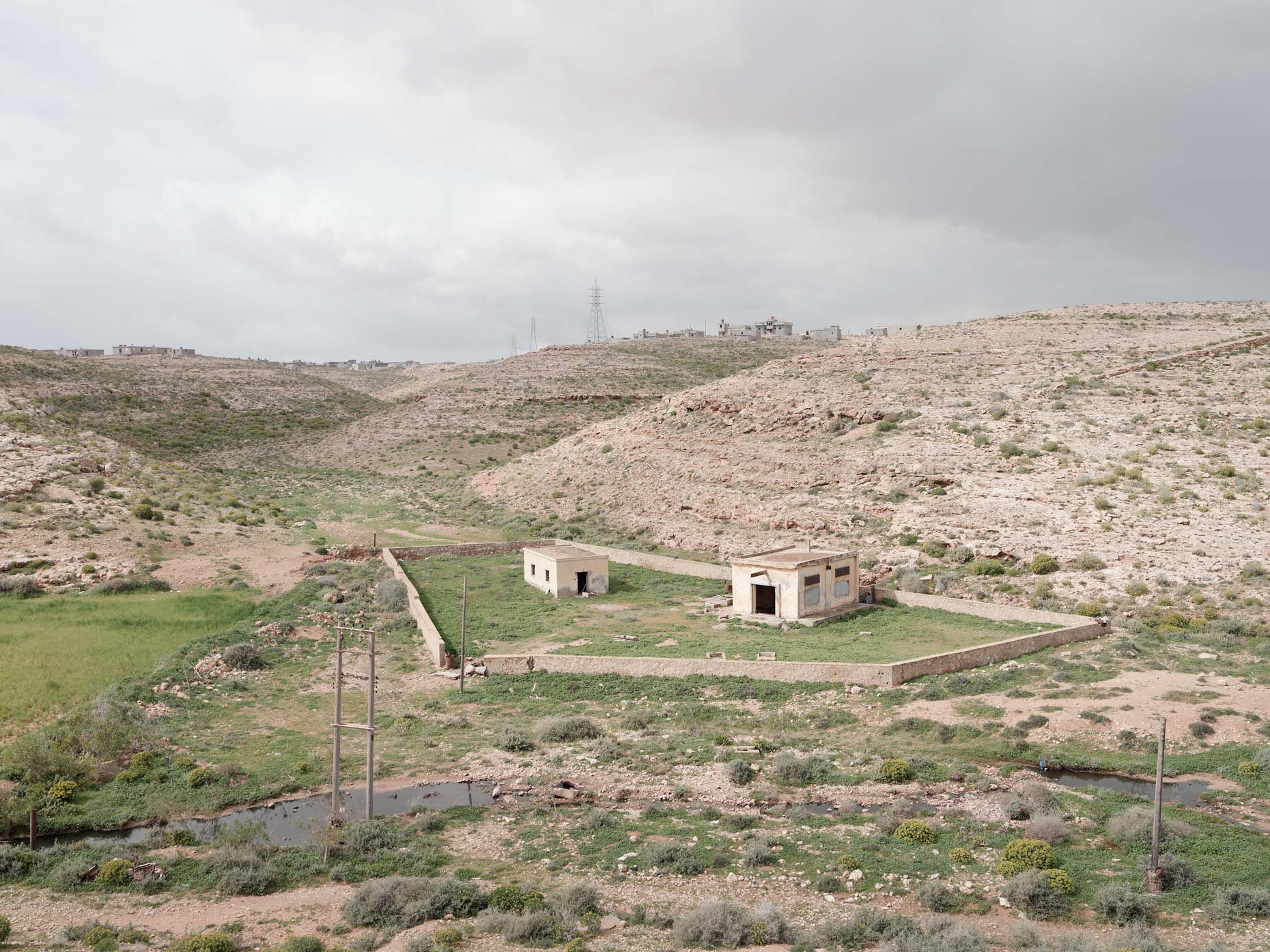 A barren desert landscape with a few small, white buildings and a green fenced area in the foreground, hills with sparse vegetation, and utility towers in the distance under a cloudy sky.