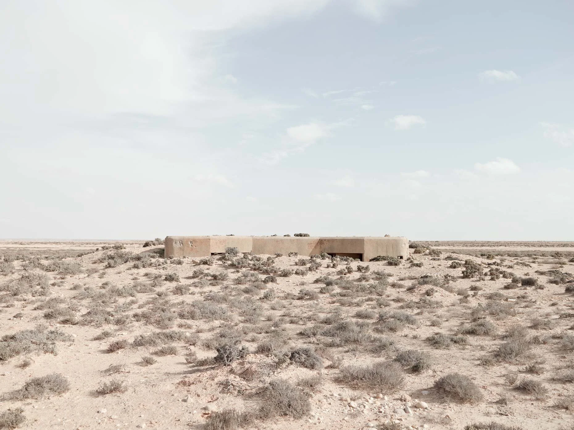 A barren desert landscape with sparse vegetation and a rectangular adobe-style building in the distance under a partly cloudy sky.