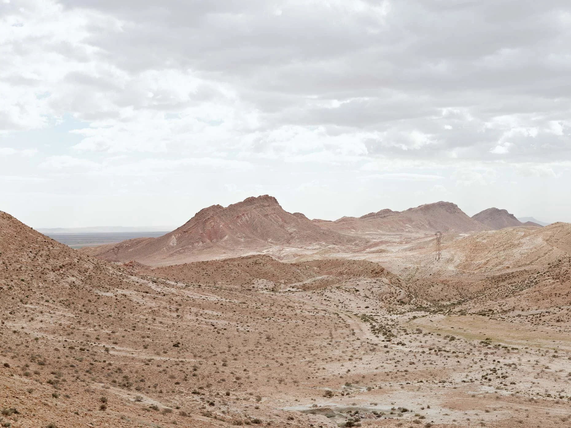 A vast desert landscape with reddish-brown, rocky hills and mountains under a cloudy gray sky.