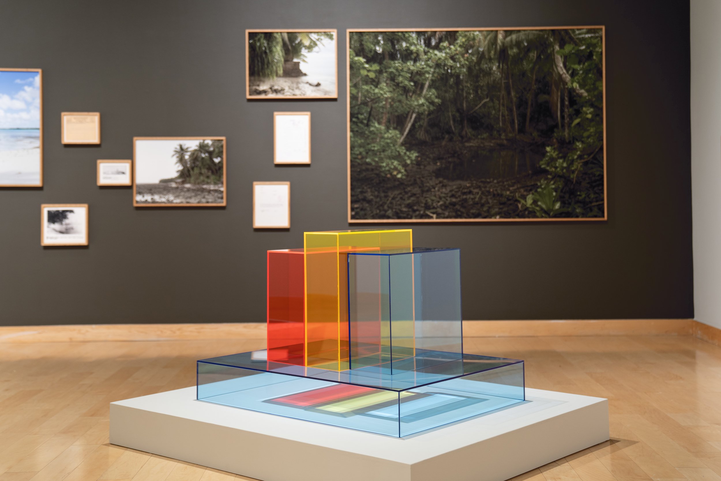 A museum display features a modern art installation of transparent colored rectangles stacked and arranged in a geometric pattern. In the background, there are framed photographs and artwork of natural landscapes and a forest scene on a dark wall.