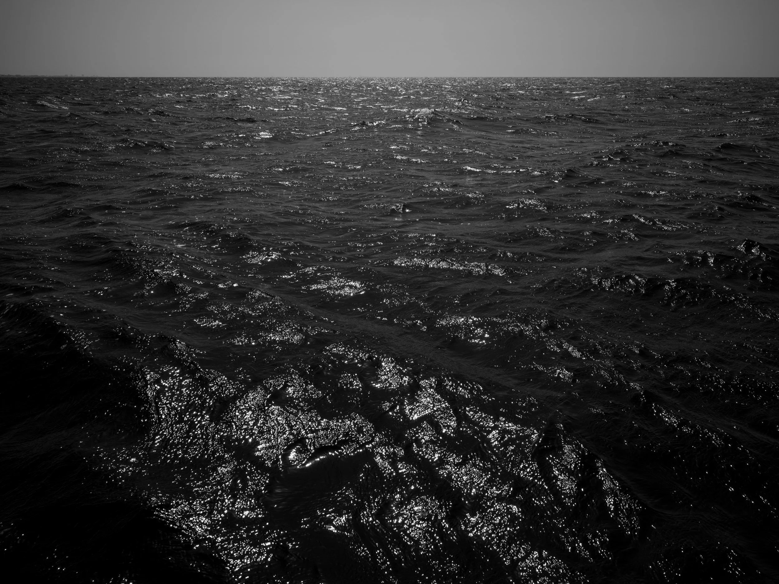 Black and white photograph of the ocean with sunlight reflecting off the surface and a distant horizon line.