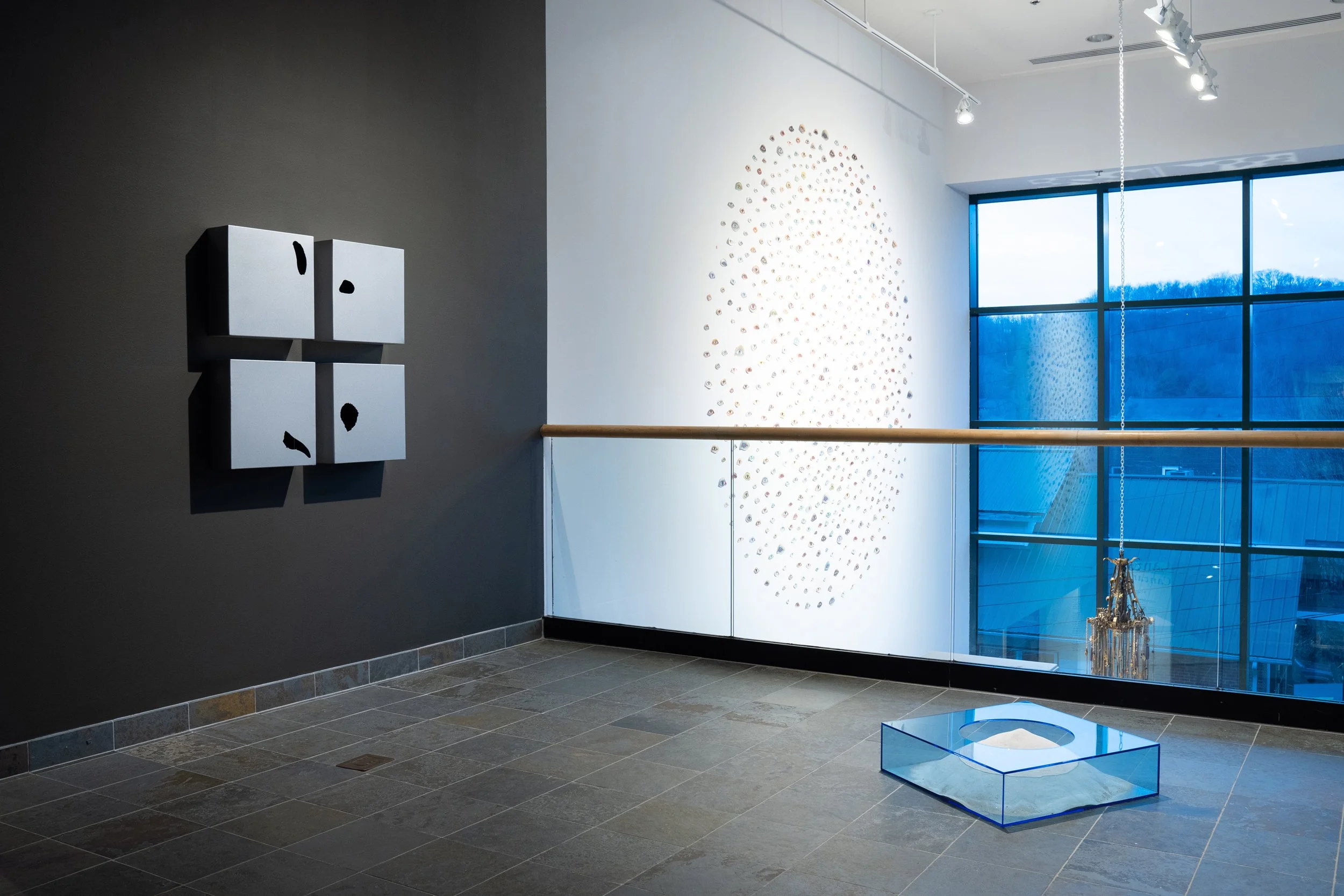 Art gallery interior with wall-mounted sculpture made of black blobs on white cubic panels, an oval tunnel-like installation with small objects on the wall, large windows showing an overcast sky, chandelier, and tiled floor.