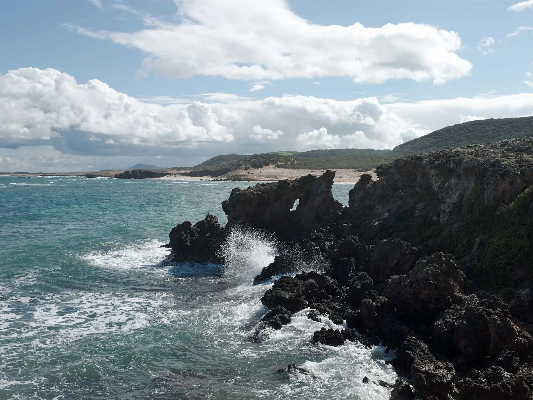 A coastal landscape with rocky cliffs in the foreground, waves crashing against the rocks, a sandy shoreline in the distance, green hills and a partly cloudy sky.