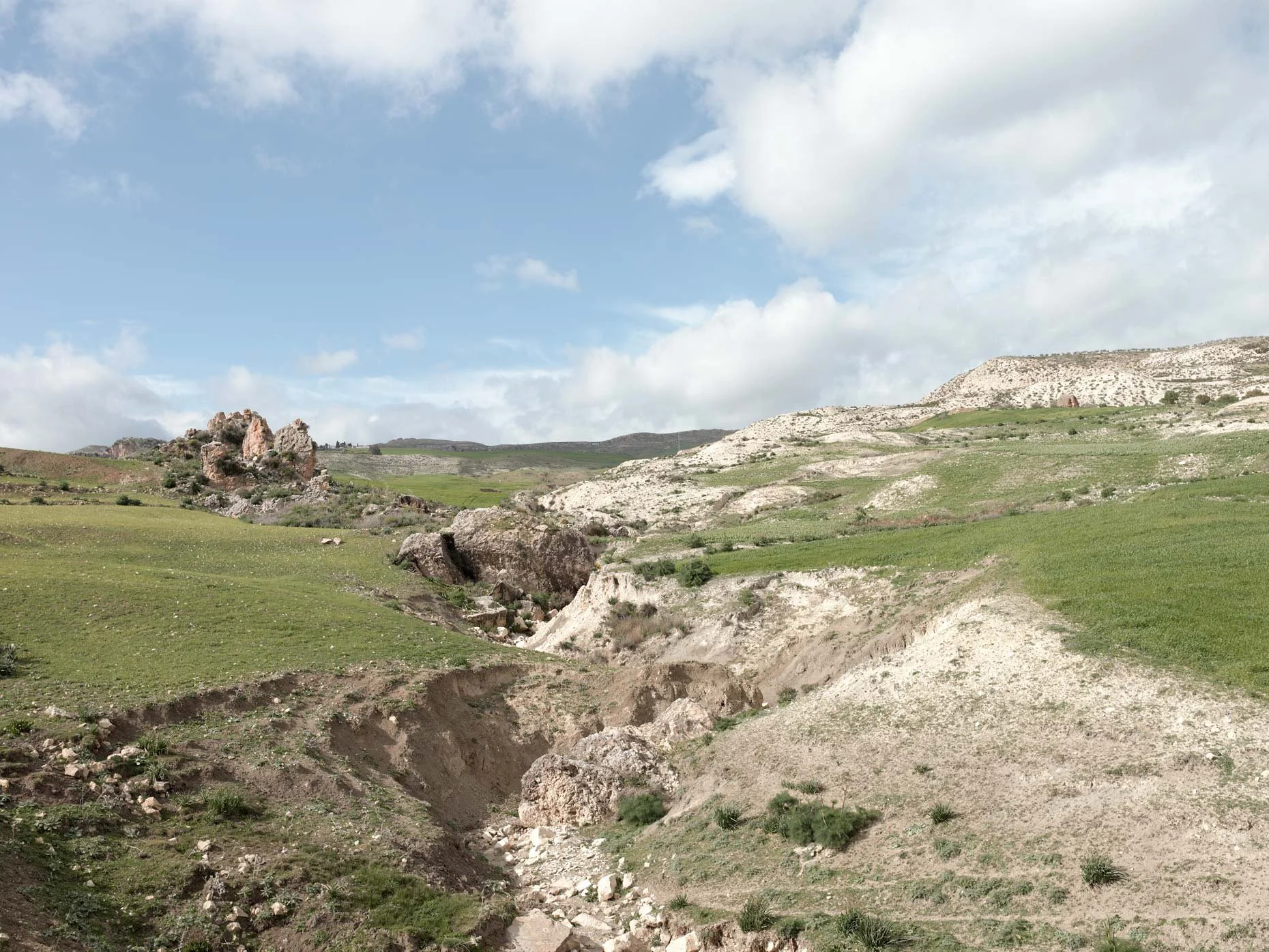 Open landscape with rolling hills, rocky outcroppings, and sparse vegetation under a partly cloudy sky.