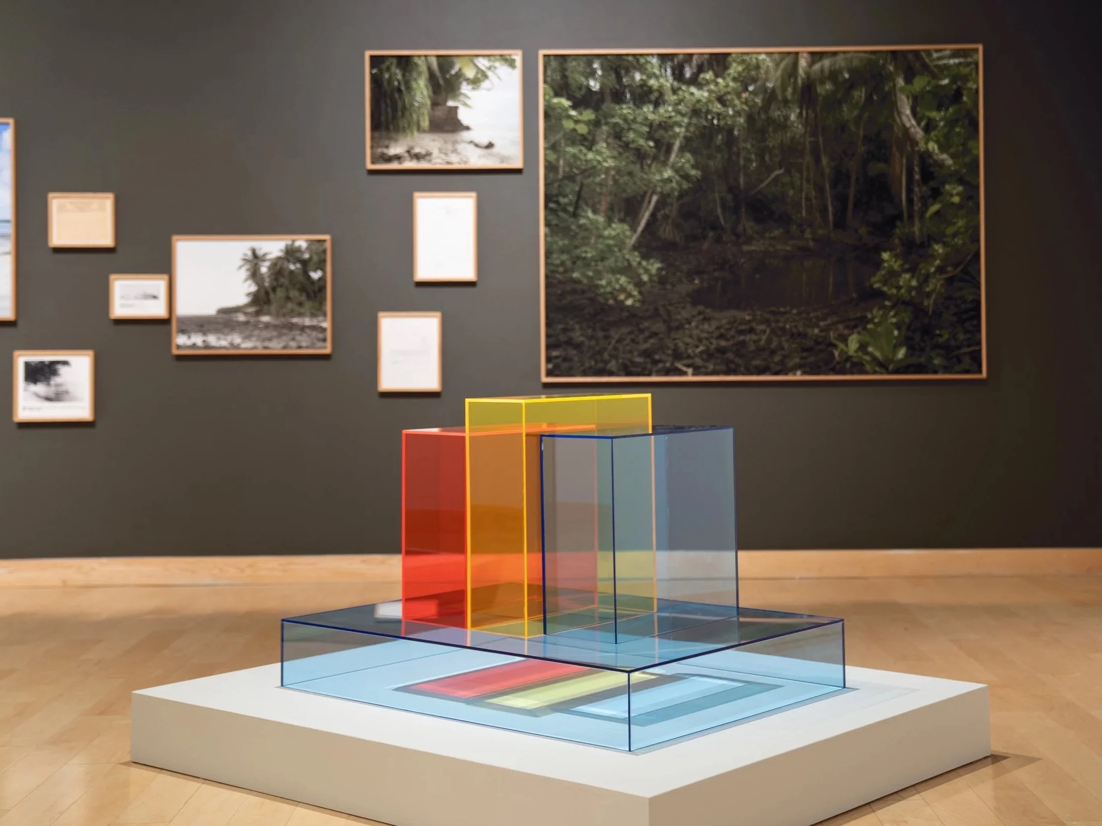 Colorful geometric glass sculpture on a white pedestal in an art gallery, with framed nature photographs on a dark wall in the background.