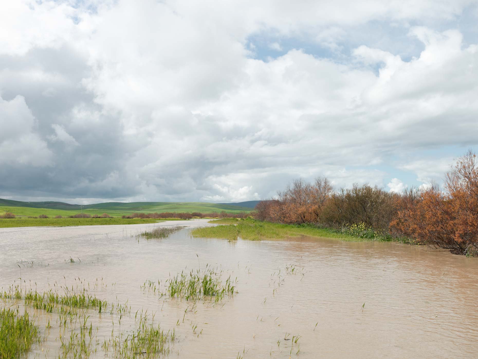 A river with surrounding green grass and trees, under a cloudy sky with patches of blue.