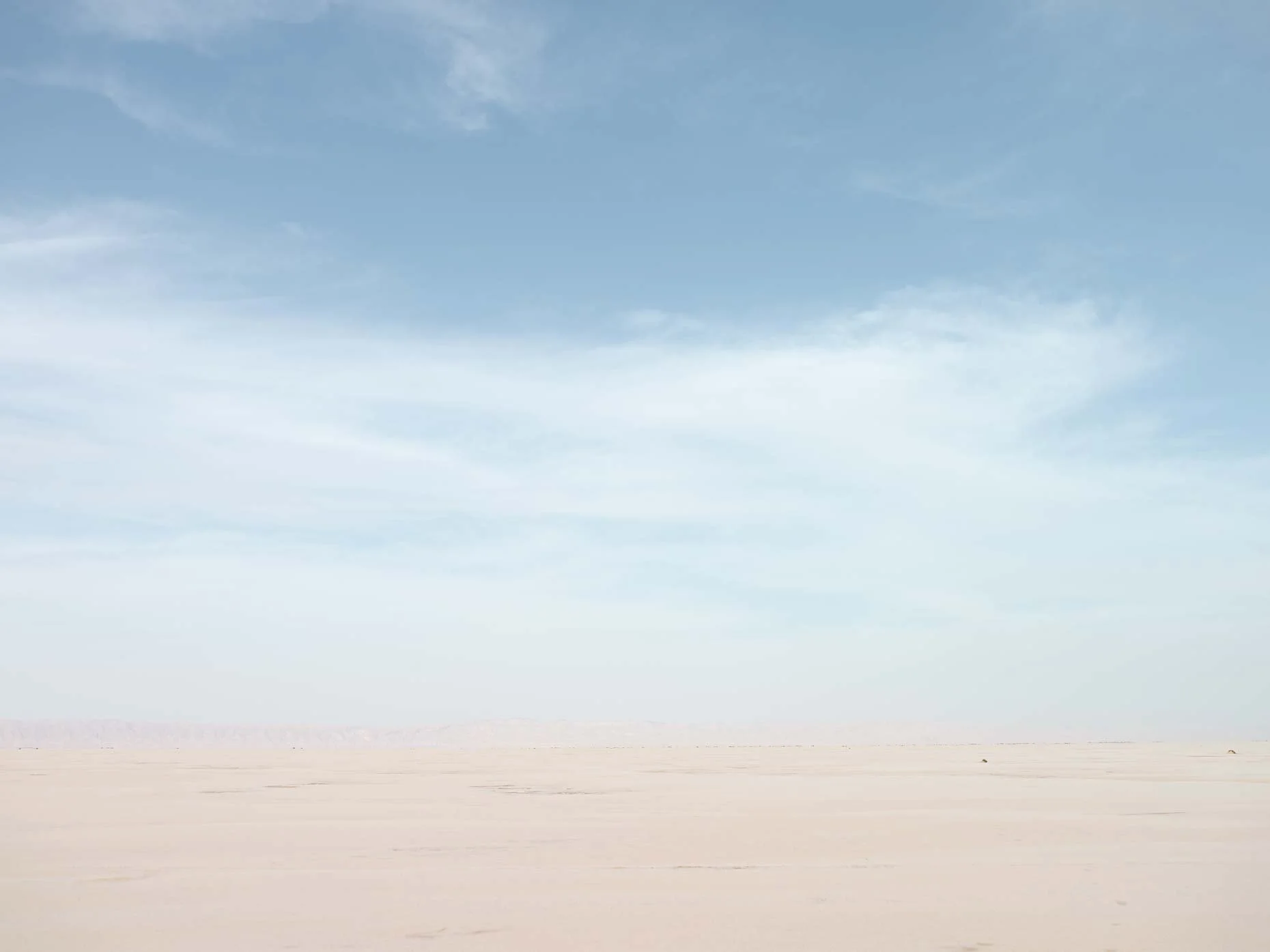 A vast flat desert landscape with a distant mountain range under a partly cloudy blue sky.