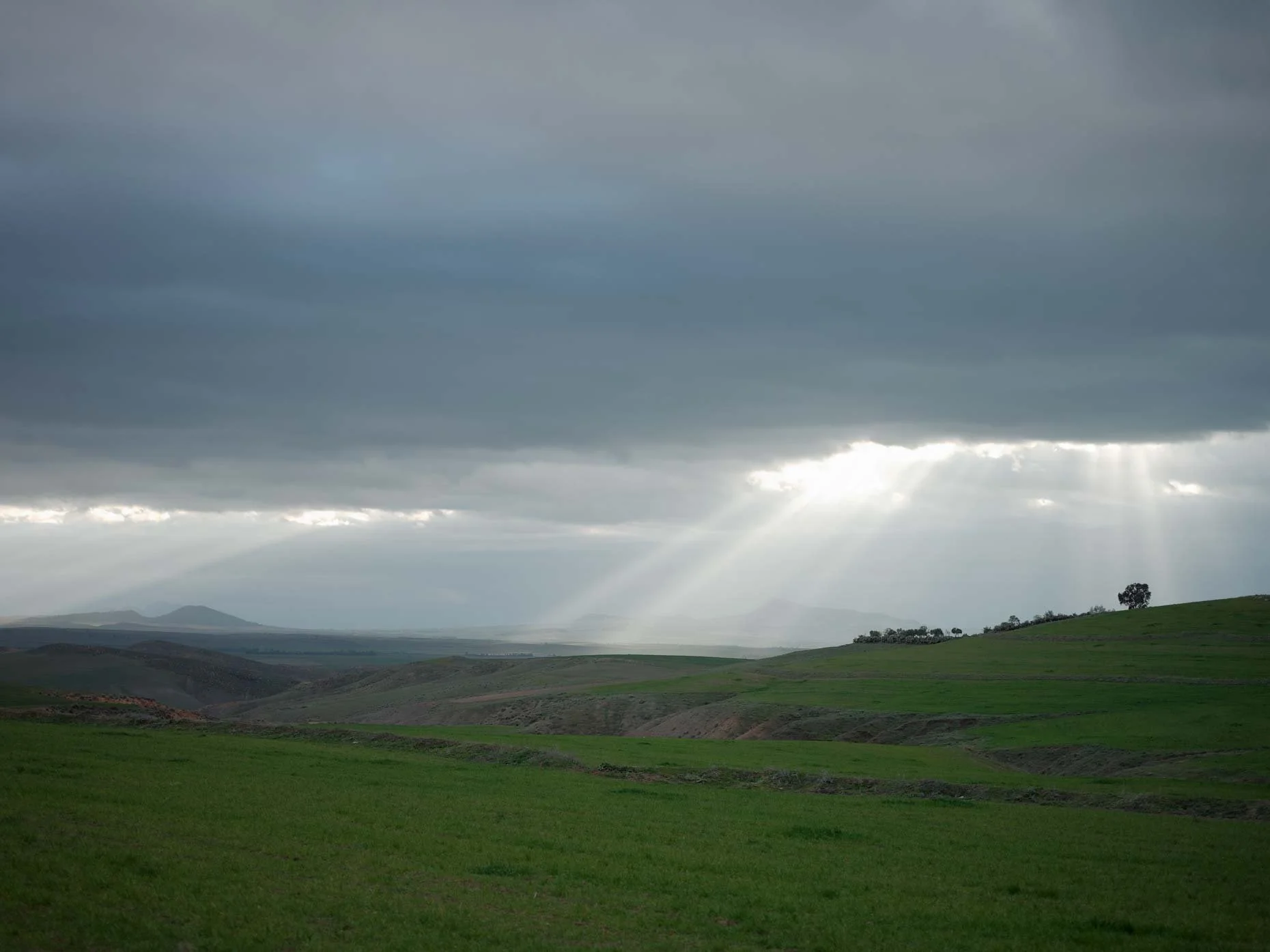 Overcast sky with sun rays breaking through clouds over a green hilly landscape.