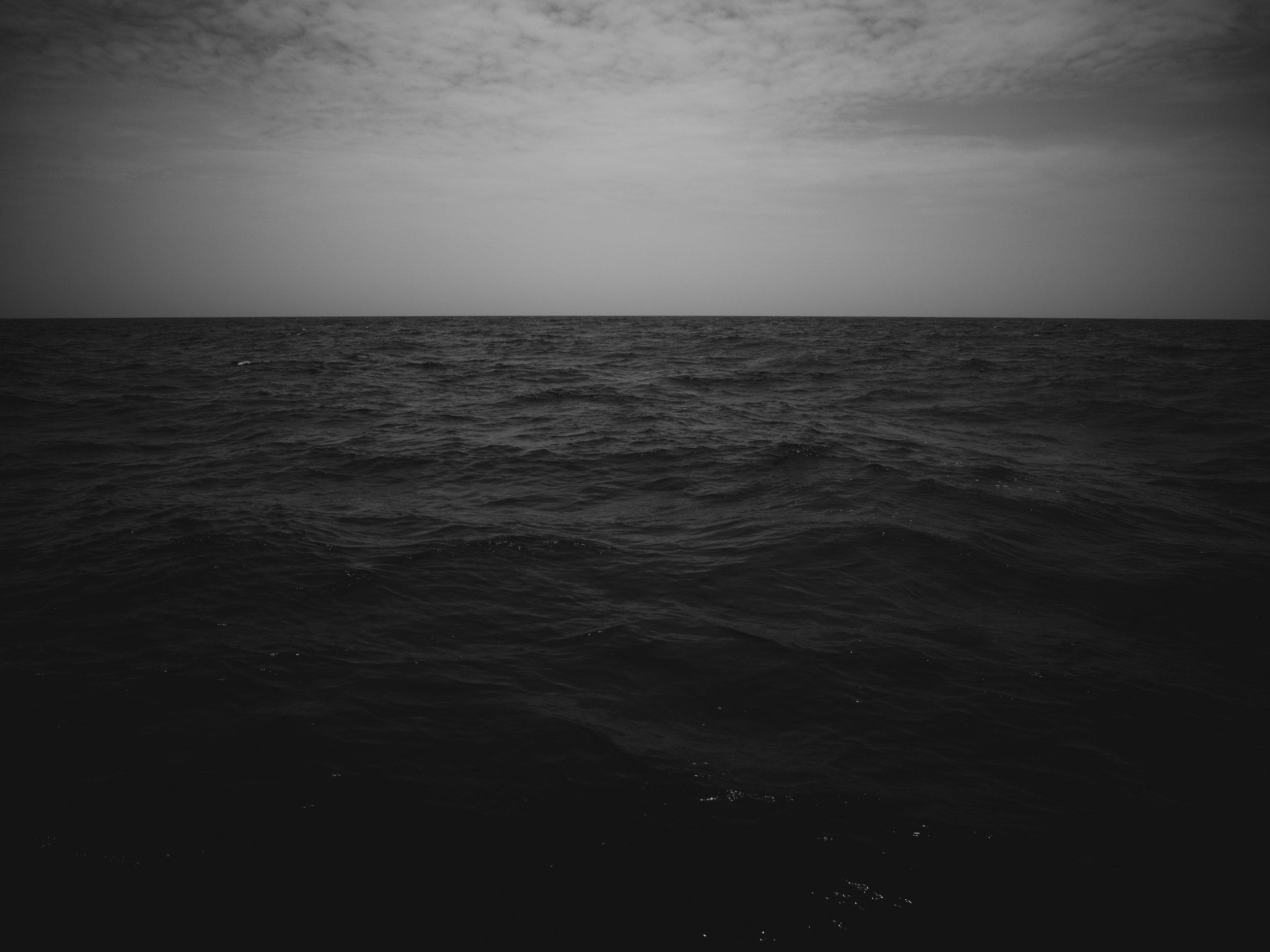 A black and white photo of the ocean with calm waves and a cloudy sky.