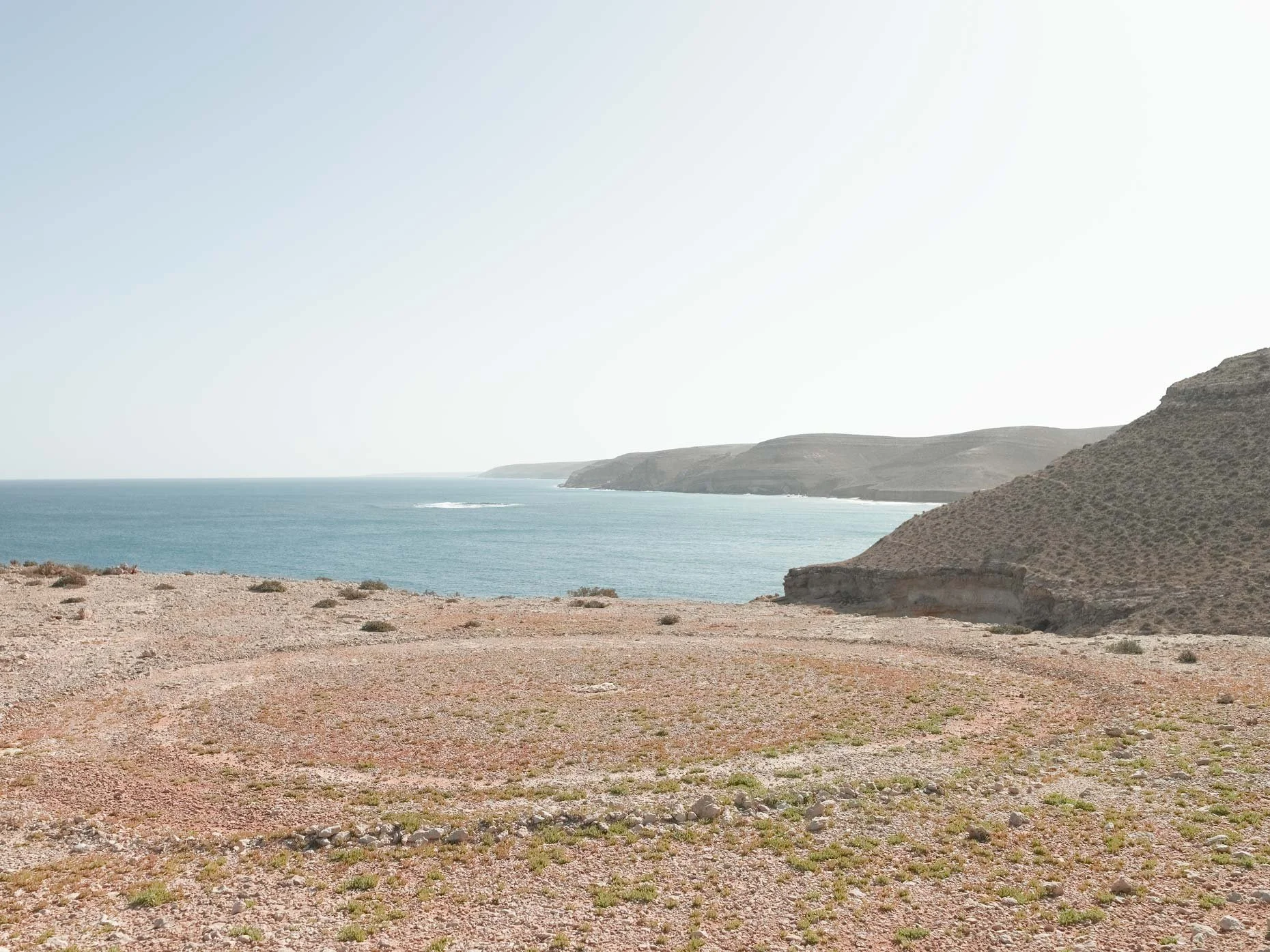 Shot of a rugged, dirt coastal landscape with sparse vegetation in the foreground, distant hills at the water's edge, and a calm ocean under a clear sky.