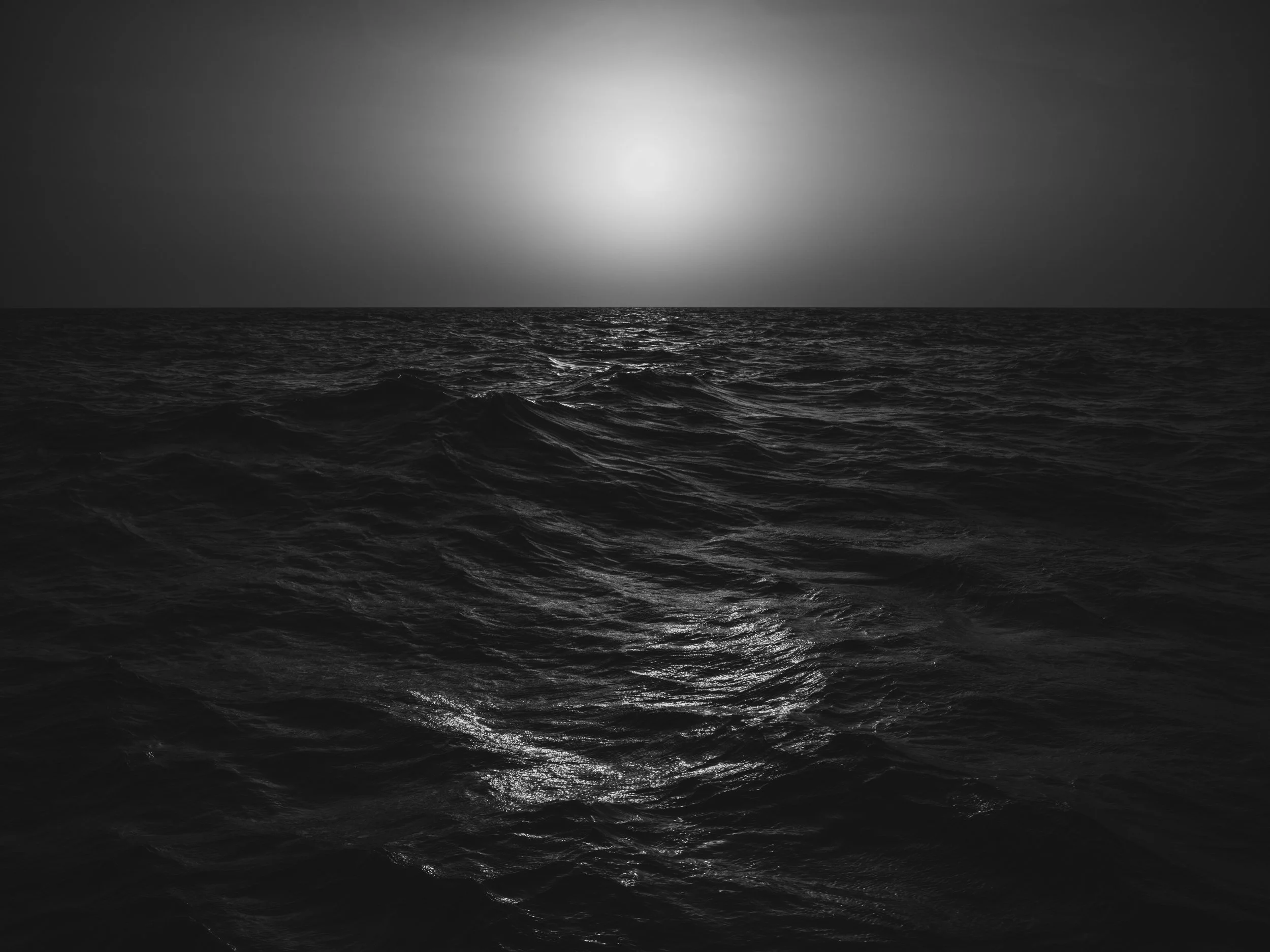 Black and white photograph of ocean waves with a bright sun reflecting on the water's surface.