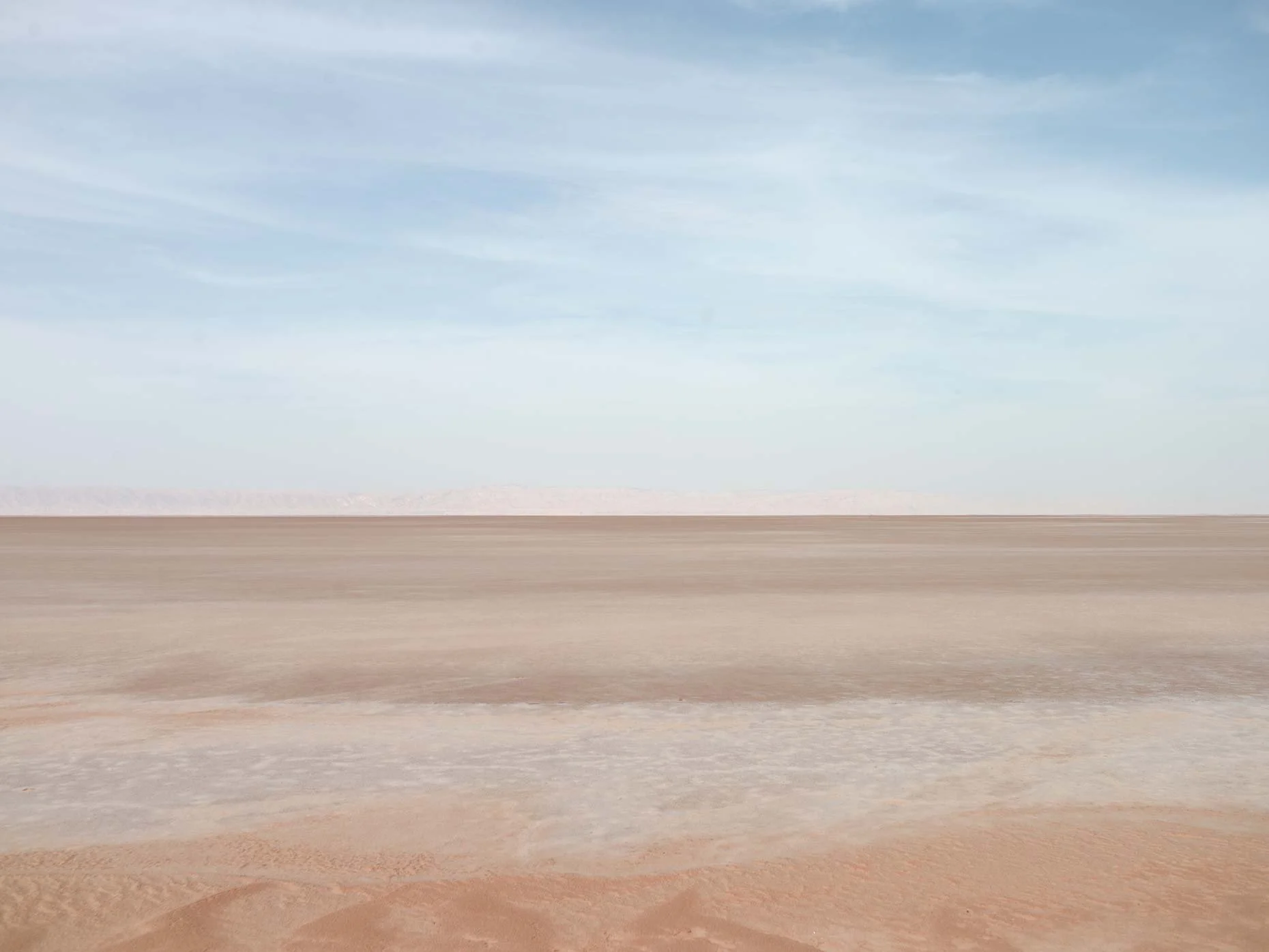 Wide view of a flat, dry desert landscape with distant mountains and a pale blue sky with wispy clouds.