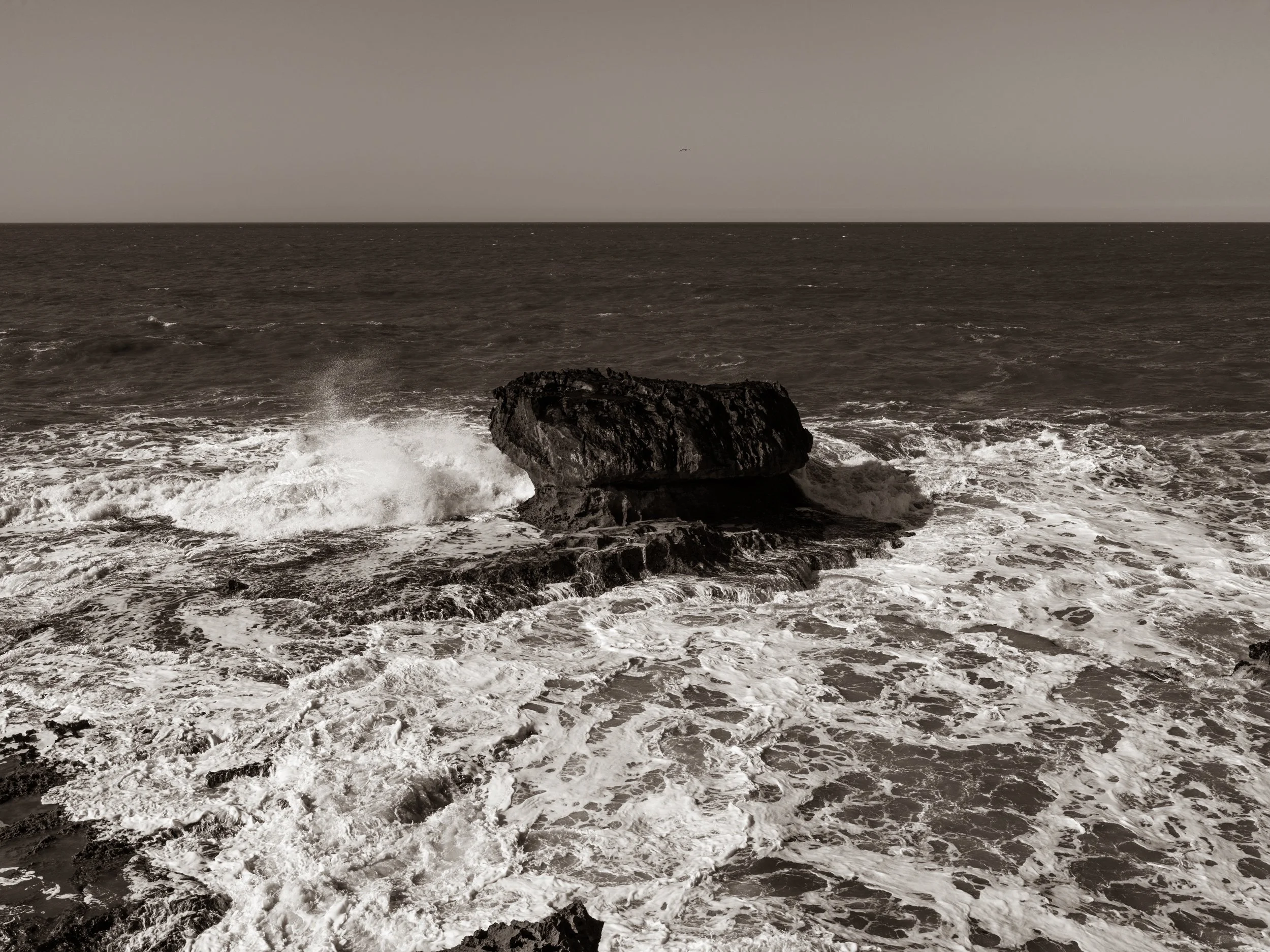 A large rock in the ocean with waves crashing against it, extending into the horizon under a cloudy sky.
