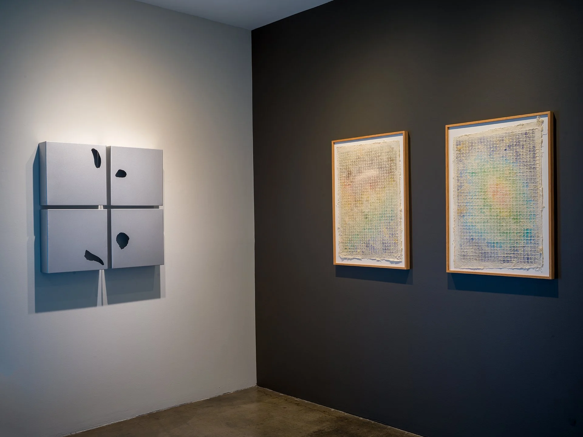 Art gallery with four framed mixed media pieces on two walls, white and dark gray, with neutral flooring.