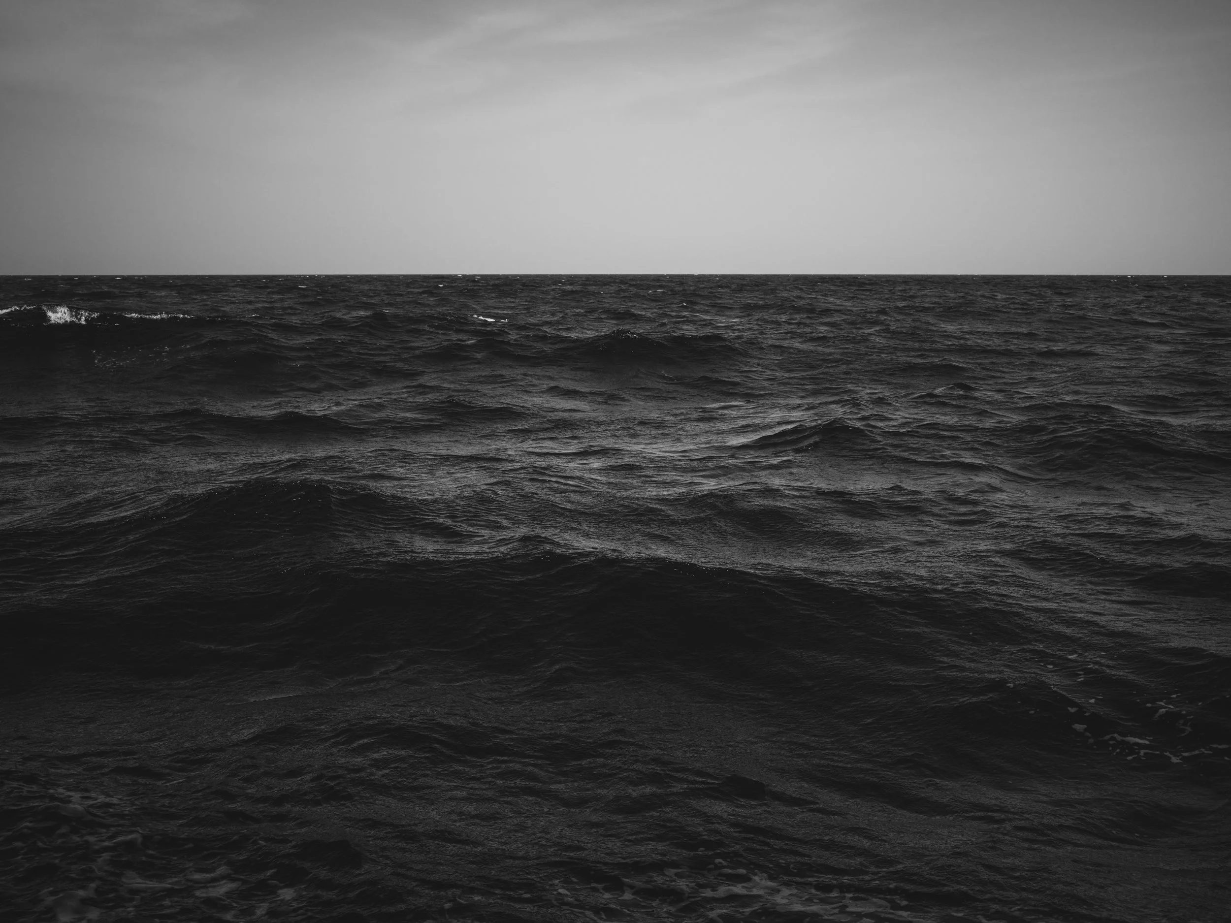 Black and white photo of the ocean with waves and a cloudy sky above.