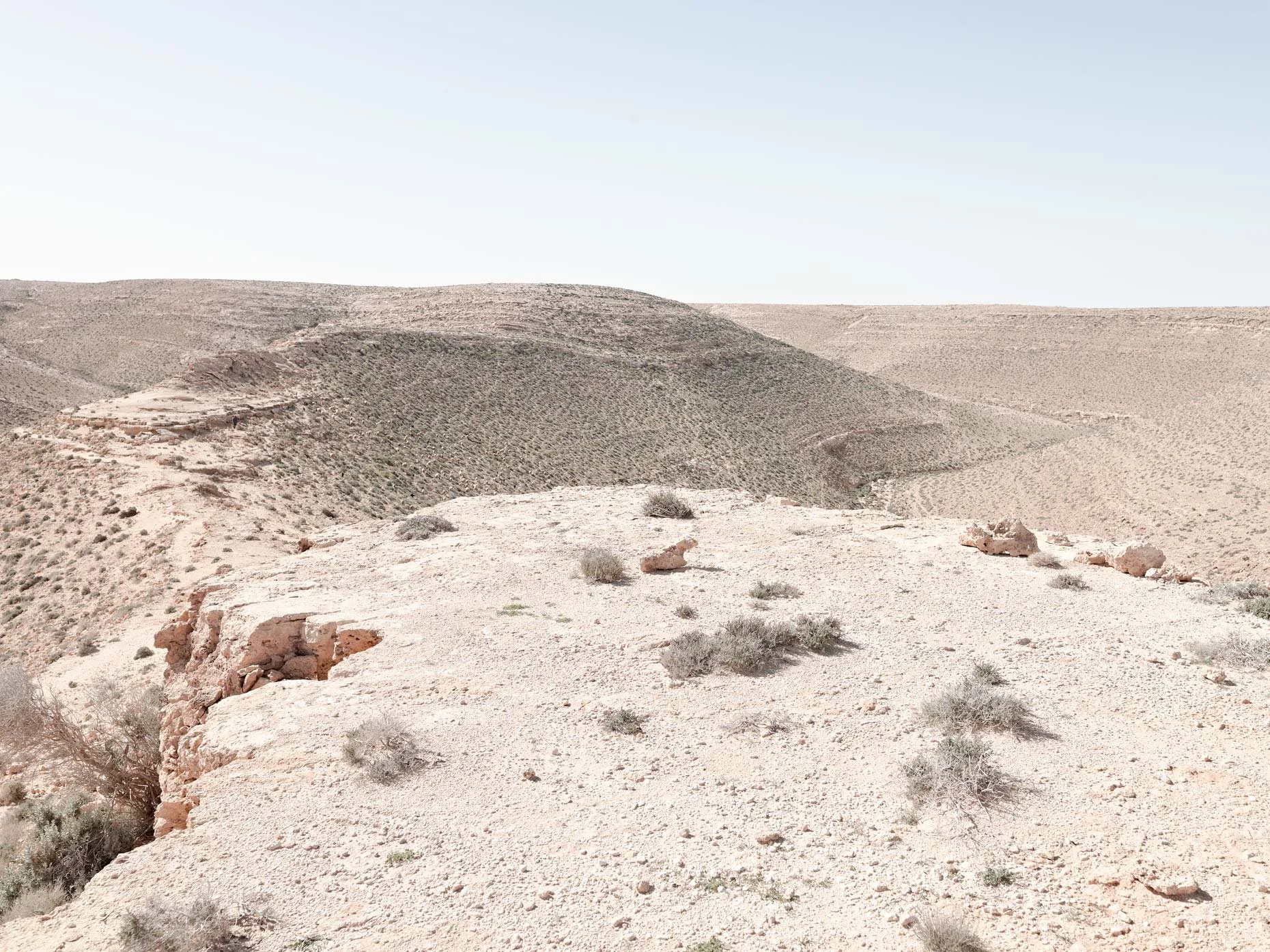 Desert landscape with rolling hills, sparse vegetation, rocks, and dry terrain under a clear sky.