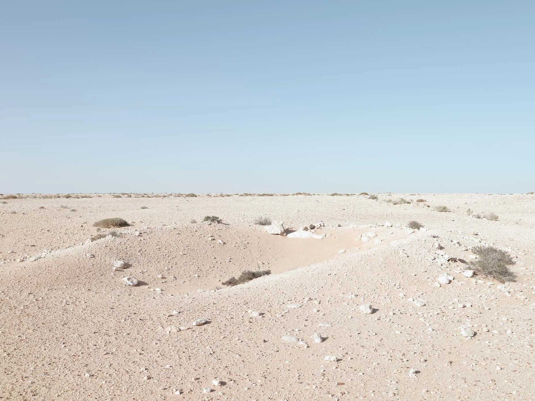 A flat, sandy desert landscape with sparse small bushes and rocks, under a clear blue sky.