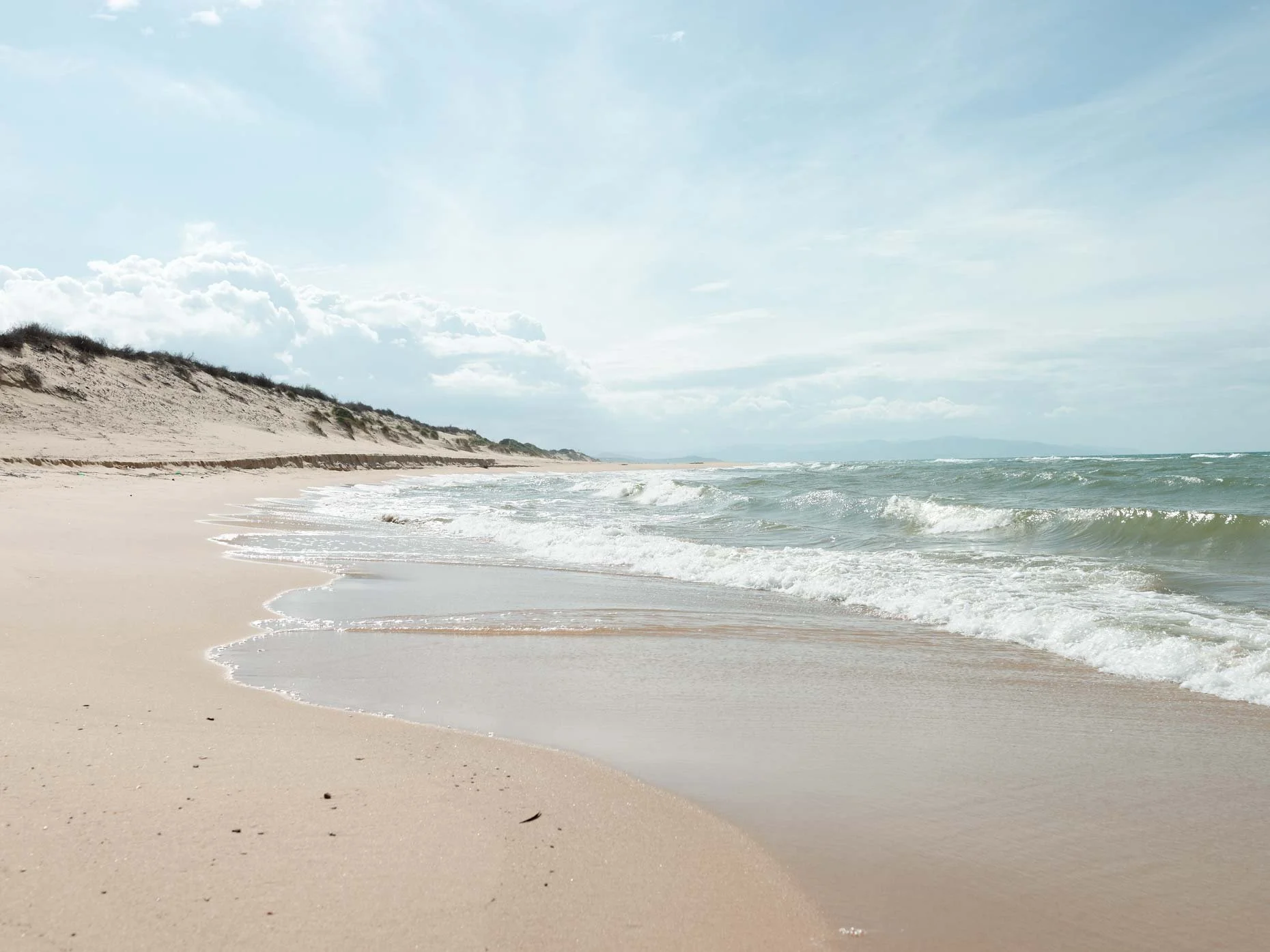 A sandy beach with gentle waves and a cloudy sky, with dunes and sparse vegetation along the coast.
