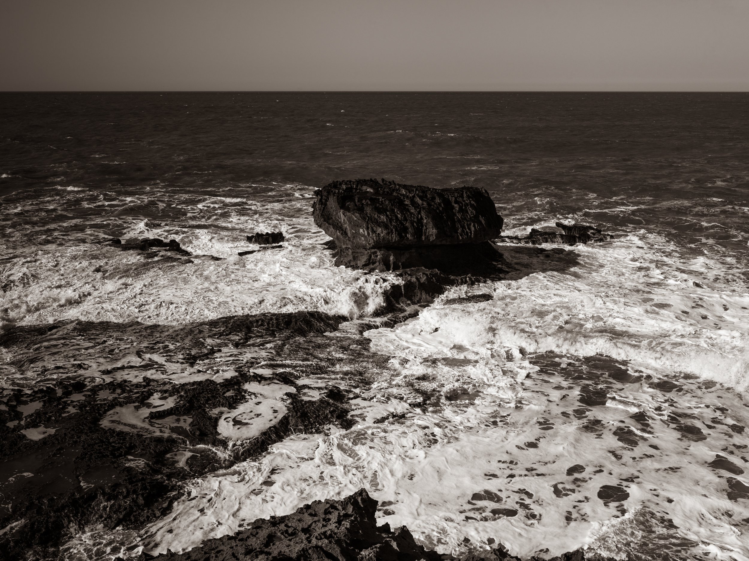 Black and white photo of ocean waves crashing against rocks on the shoreline with a distant, calm sea and cloudy sky.