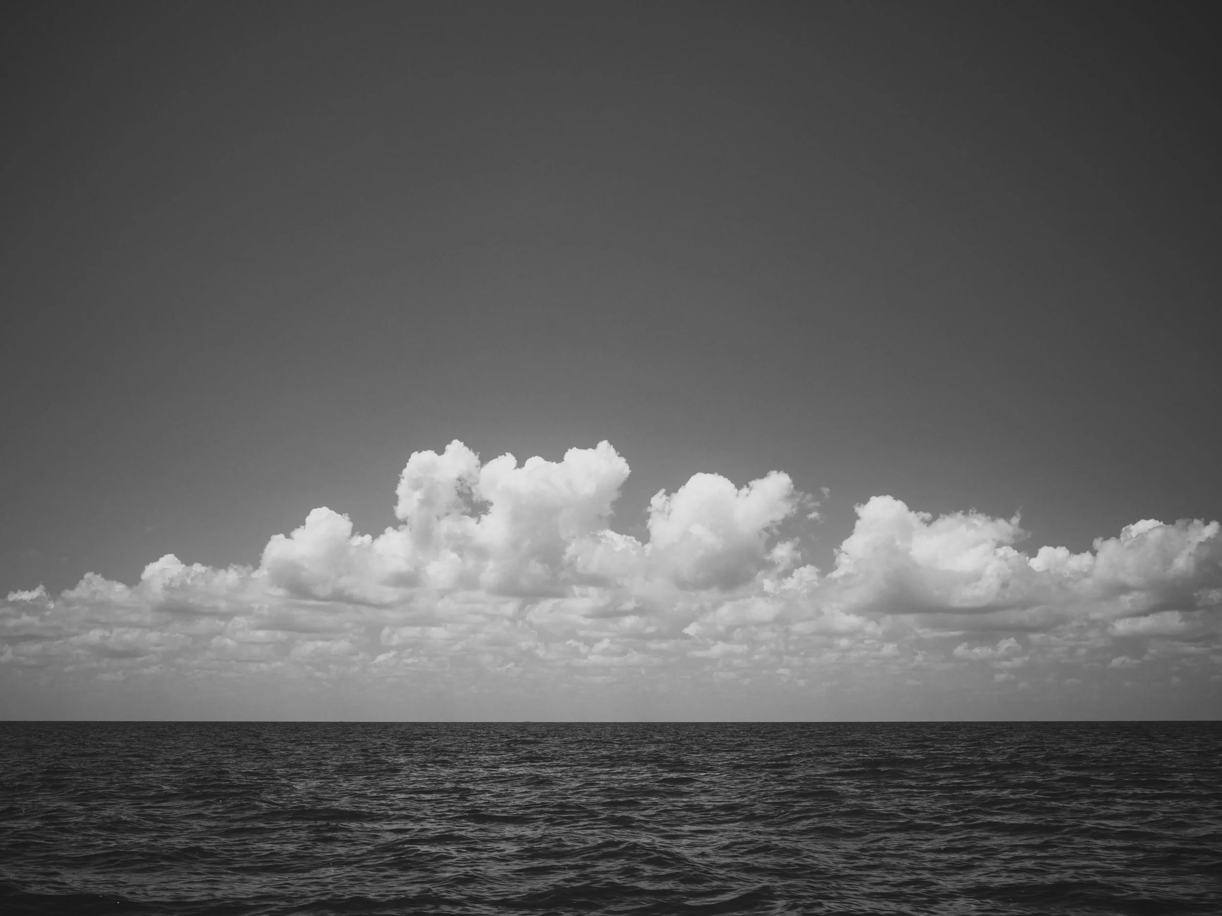 Black and white photo of cloud-filled sky over a dark ocean horizon.