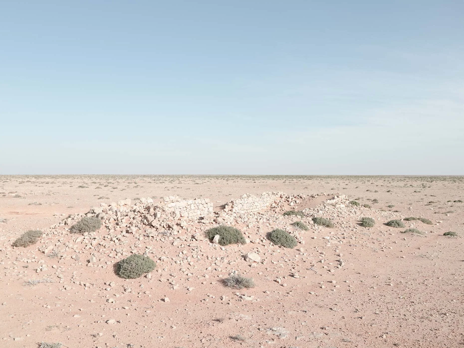 A vast desert landscape with sandy terrain and sparse green bushes, under a clear blue sky with some light clouds.