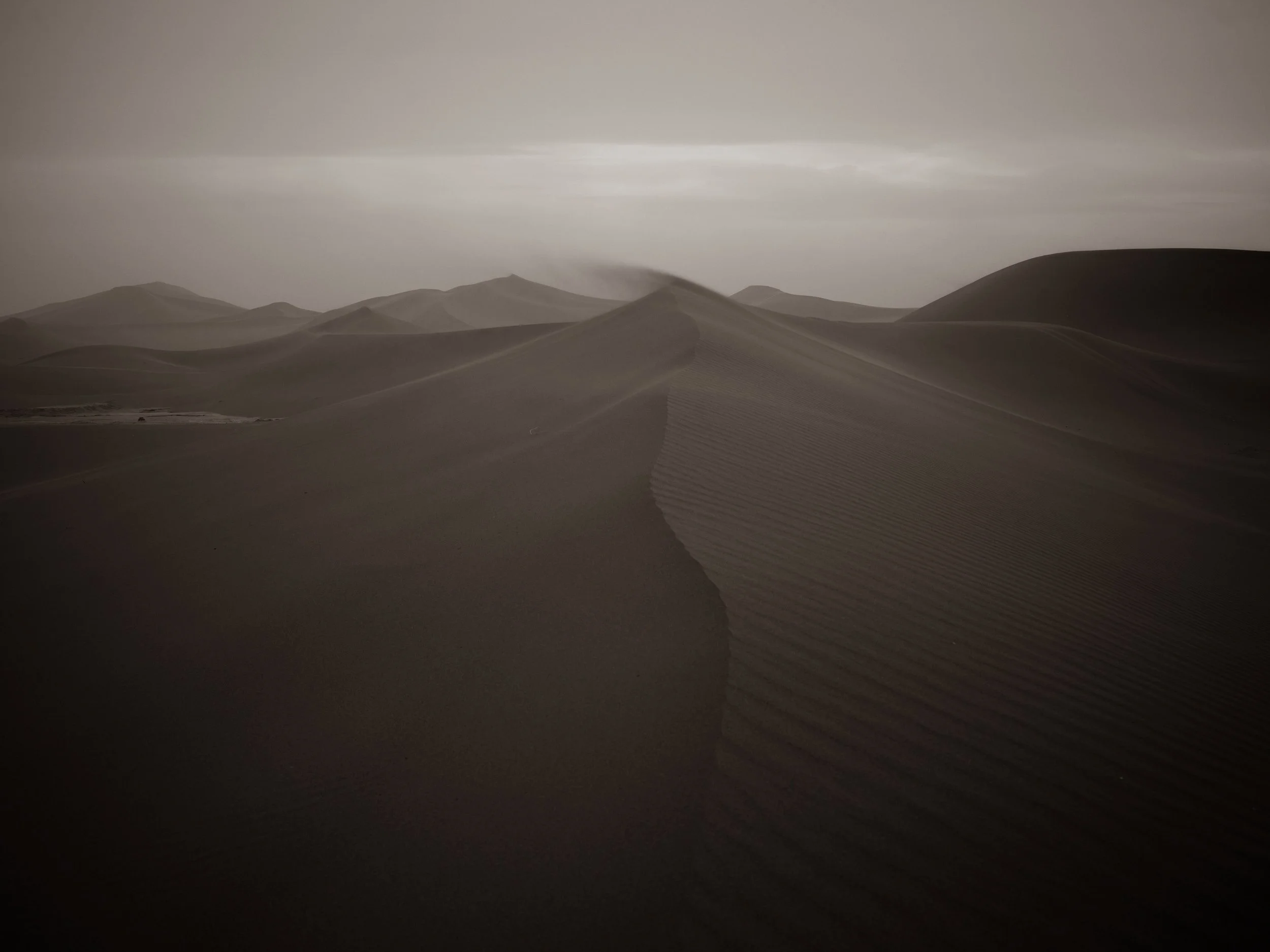 Photo of sand dunes in a desert with smooth, sweeping curves and layered ridges under a cloudy sky.