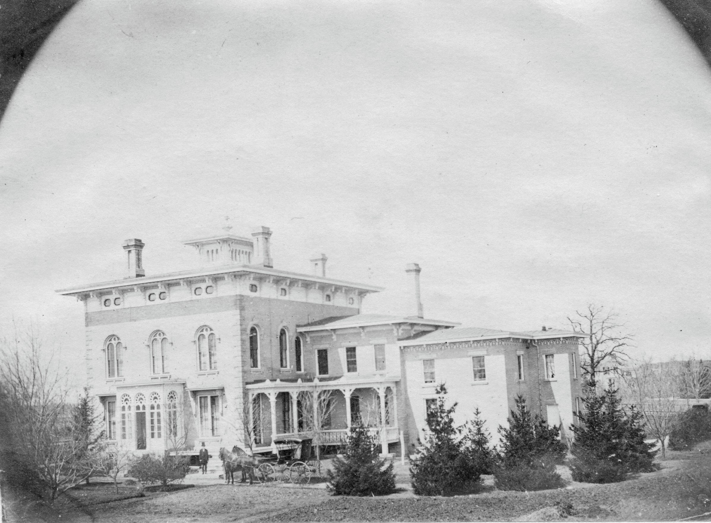 A black and white photo of a large, ornate Victorian-style house with multiple gables, turrets, and balconies. The house is surrounded by trees and a lawn with a few people and horse-drawn carriages nearby.
