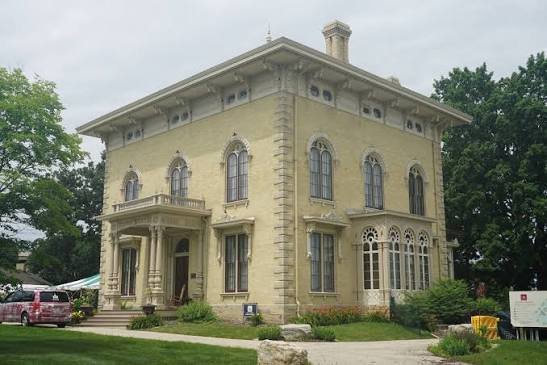 A historic, two-story yellow mansion with large windows, decorative architectural details, and a small front porch, surrounded by green trees and a well-maintained lawn.