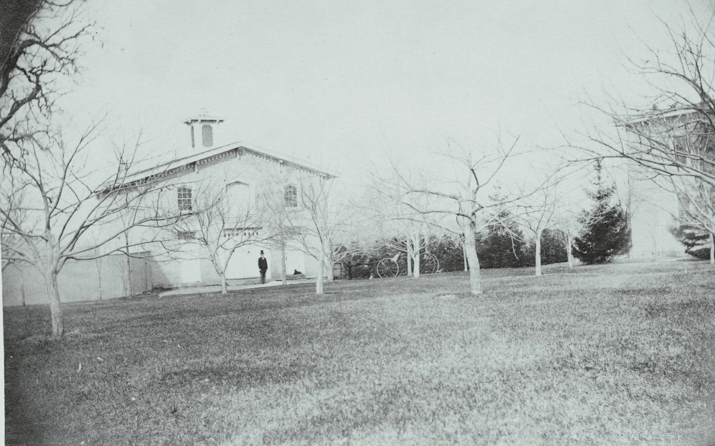 A black and white photo of a house surrounded by leafless trees, with a person standing near the house, and bicycles leaning against the wall.