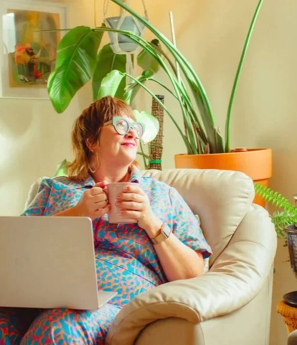 A woman with short brown hair, glasses, and earrings sits on a beige armchair, holding a pink mug. She has a laptop on her lap and appears relaxed with a cheerful expression. Large green houseplants behind her and a colorful painting on the wall.