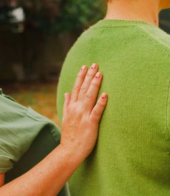 A person places their hand on the shoulder of another person wearing a green sweater in an outdoor setting.