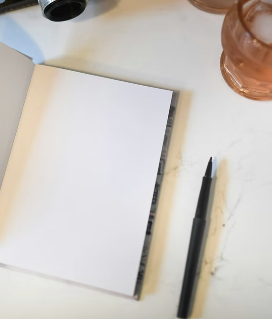 Open notebook with blank white page on a white surface, black marker pen beside it, and glasses with a copper-colored frame partially visible, along with a black bottle and a glass of pink-colored beverage.