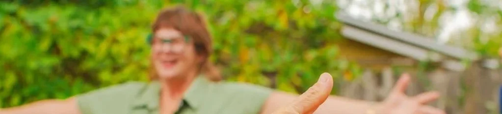 Person outdoors with arms outstretched, smiling, blurry background of green foliage and a house.