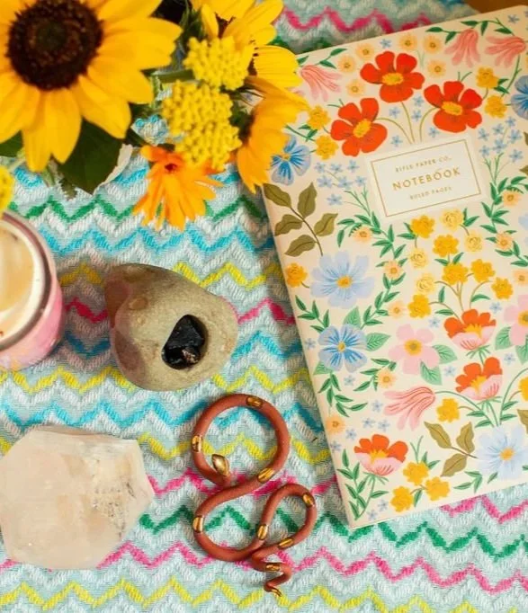 Sunflowers and yellow flowers in a vase, a colorful floral notebook, a heart-shaped rock, a solid-colored crystal, a pink and gold bracelet, and a geode on a rainbow-colored zigzag-patterned cloth.