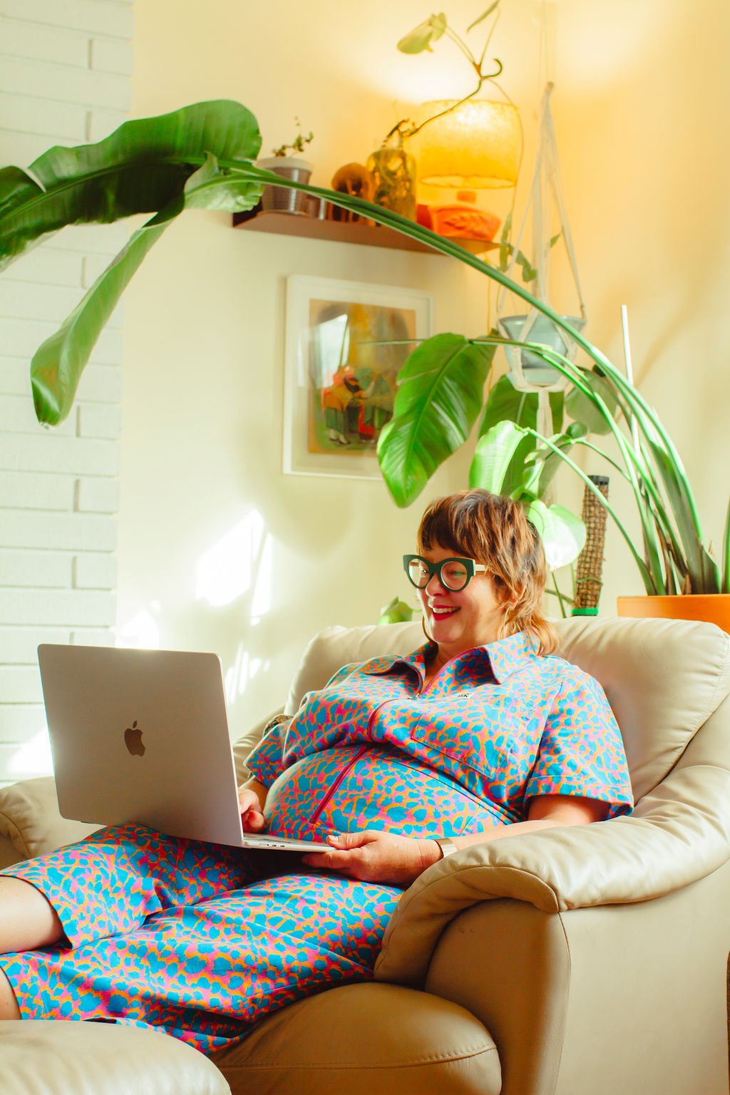 A person with curly hair, glasses, and a big smile, sitting on a beige couch and using a MacBook laptop, in a room with large green tropical plants, artwork on the wall, and warm lighting.