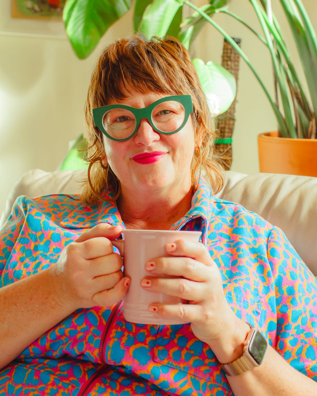 A woman with short reddish hair wearing large green glasses, a colorful animal print top, and a smartwatch, sitting on a light-colored sofa while holding a pink mug, with large green leafy plants in the background.