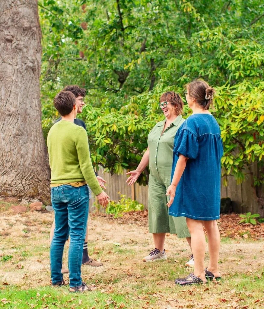 Four people standing outdoors in a garden, engaged in conversation and laughter, surrounded by green trees.