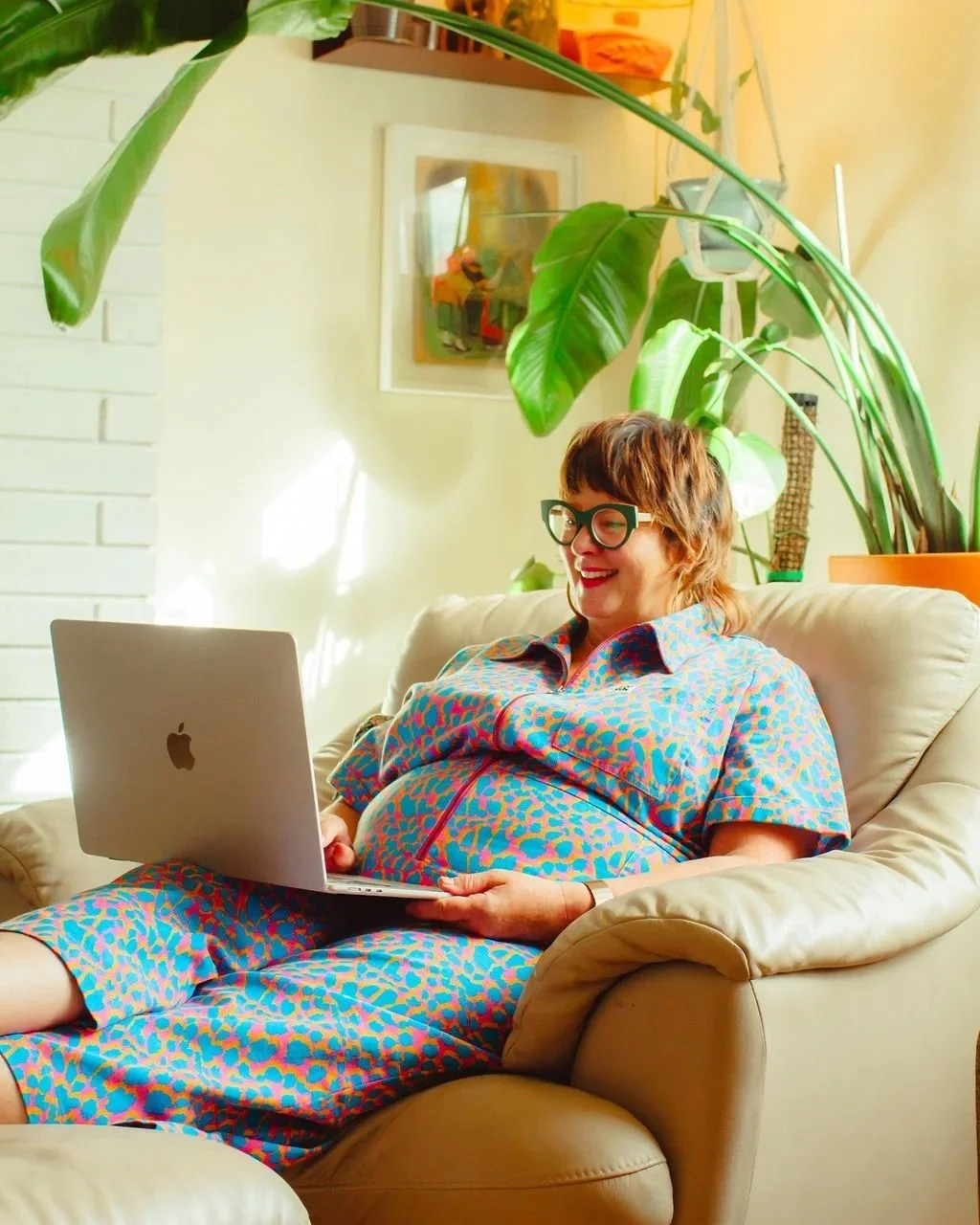 A smiling person with short brown hair, wearing large glasses and a colorful animal print dress, sitting on a beige couch with a laptop on her lap in a bright living room with greenery and artwork.