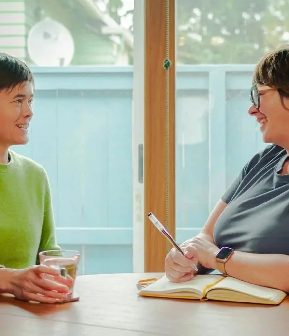 Two women sitting at a wooden table having a conversation indoors, with a sliding glass door and a backyard visible in the background. One woman wearing a green sweater holds a glass of water, while the other woman with glasses and a smartwatch is writing in a notebook.