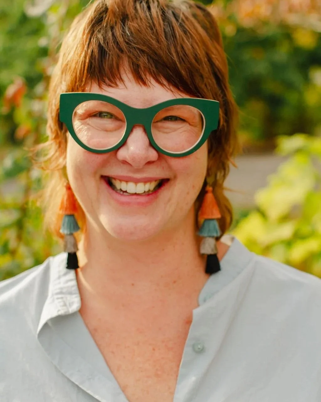 A cheerful woman with red hair wearing green glasses, orange and turquoise earrings, and a light-colored shirt outdoors with greenery in the background.