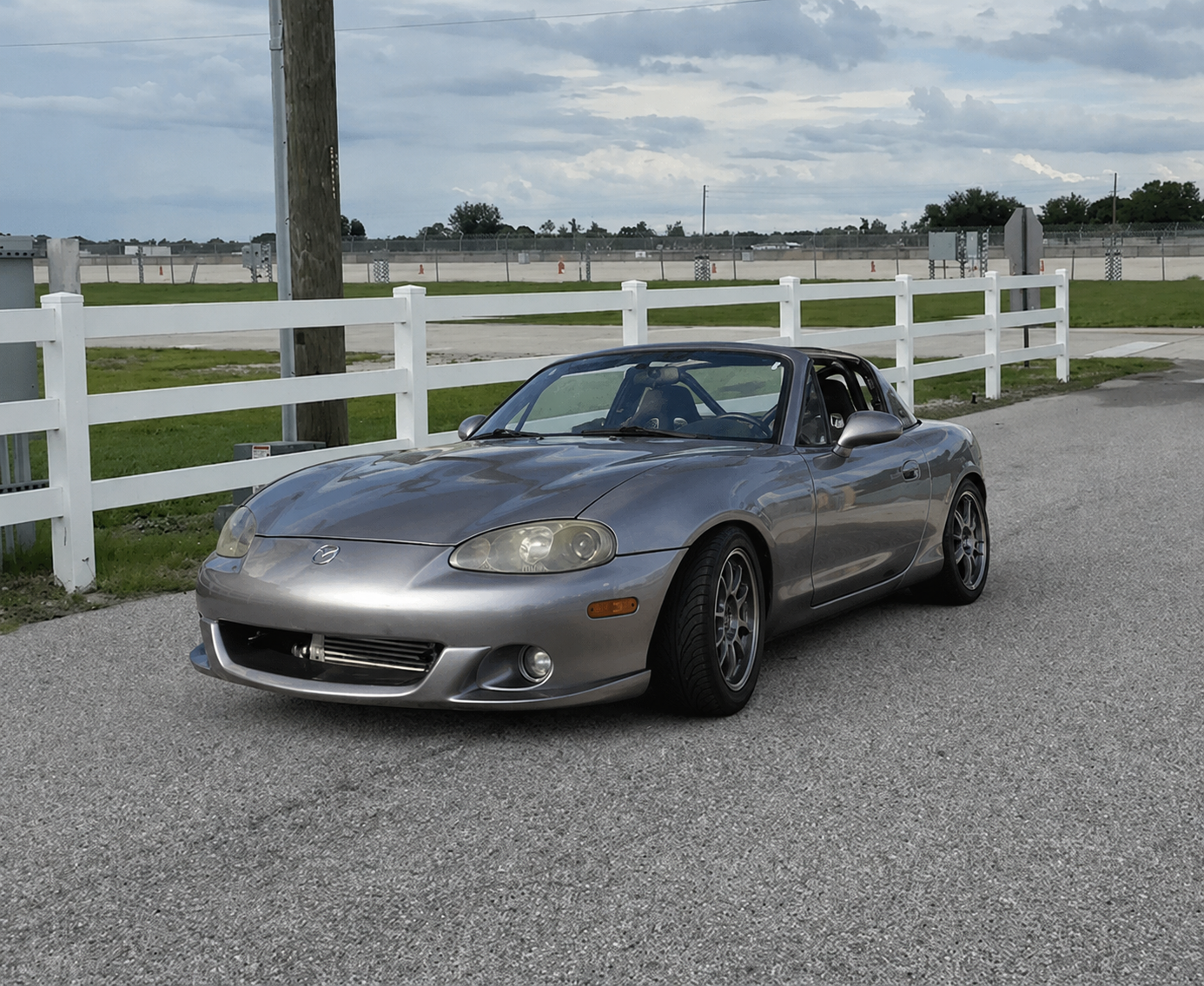 Silver Mazda MX-5 Miata convertible parked on a paved surface near a white wooden fence with green grass and cloudy sky in the background.